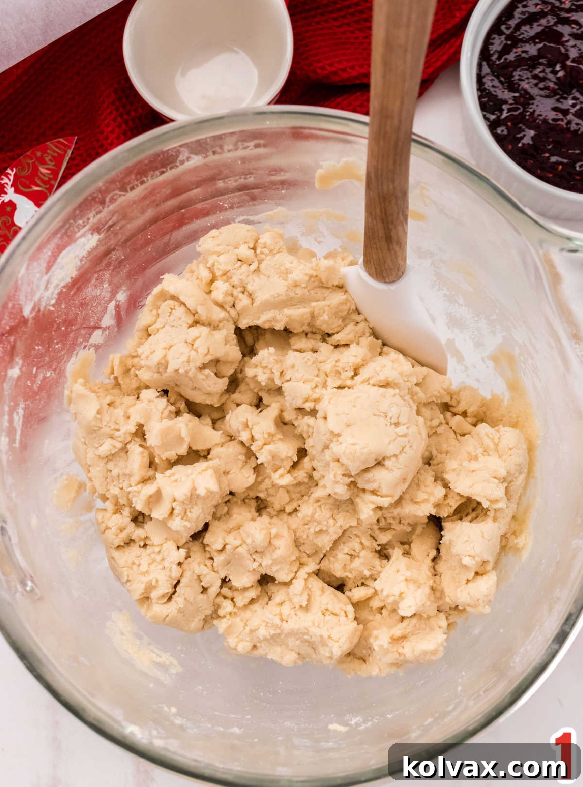 Shortbread cookie dough ready for baking in a clear mixing bowl, with a white spatula resting nearby and a bowl of bright red jam in the soft background.