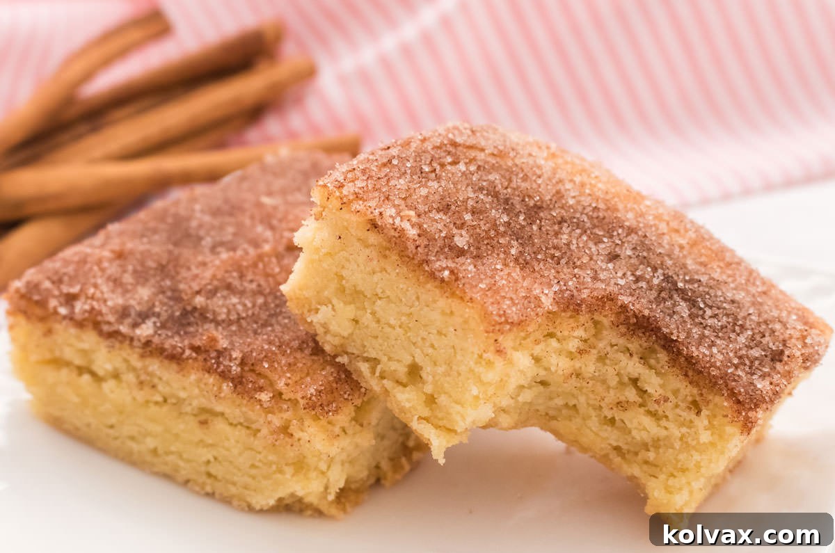 Two Snickerdoodle Bars on a white table with cinnamon sticks and a pink kitchen towel in the background.