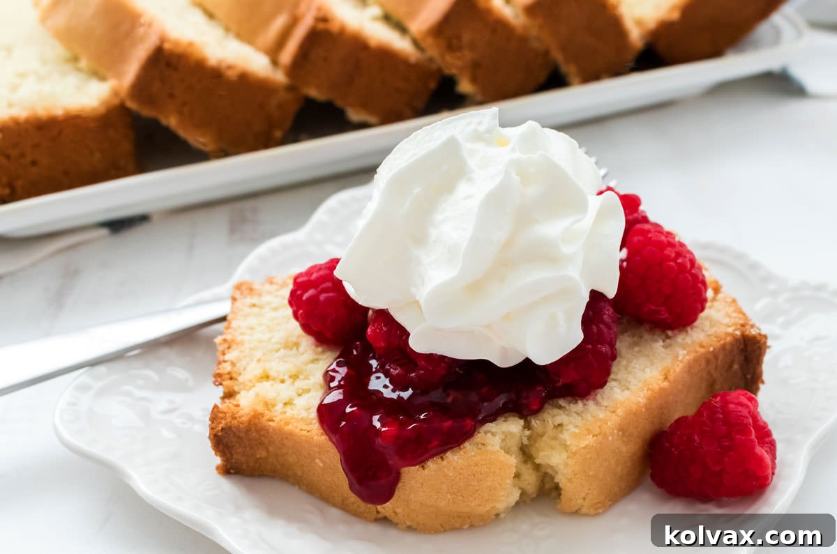 A generous slice of golden-brown classic pound cake, adorned with vibrant fresh raspberries and a dollop of fluffy whipped cream, resting on a white ceramic plate. The rest of the beautifully baked loaf is visible in the soft-focus background.