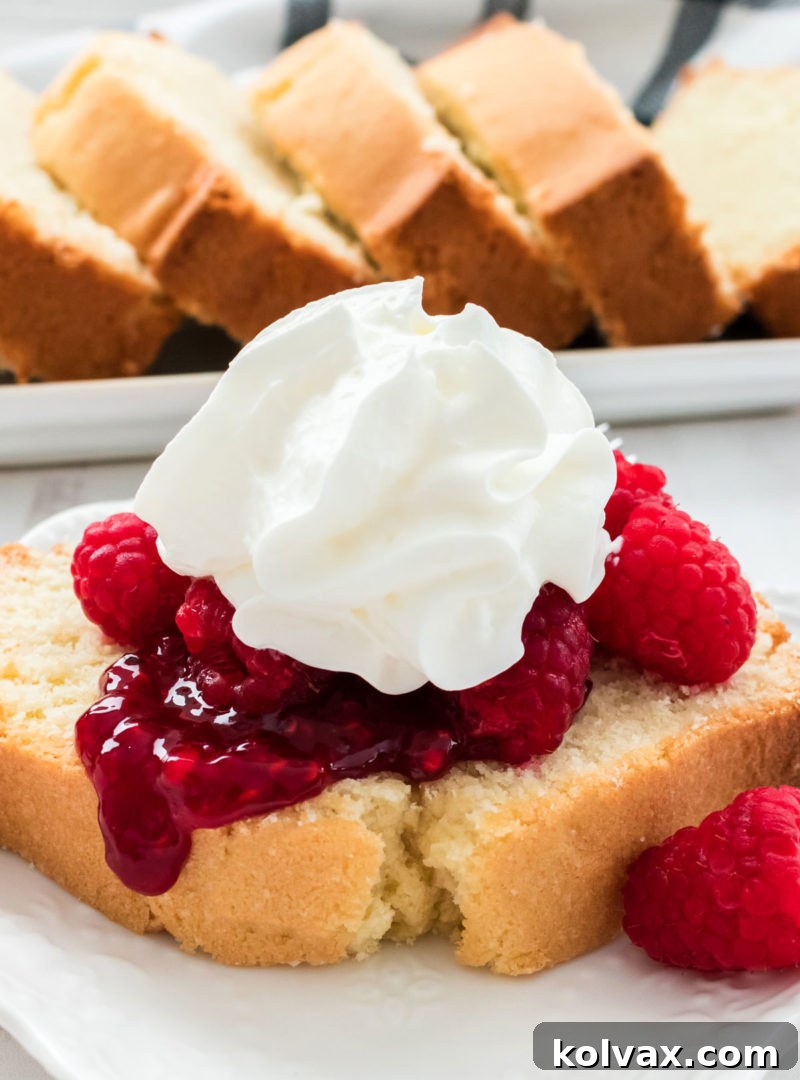 A close-up view of a perfectly baked slice of Classic Pound Cake, elegantly topped with fresh, vibrant raspberries and a dollop of airy whipped cream. Additional slices of pound cake are visible in the soft background, inviting indulgence.
