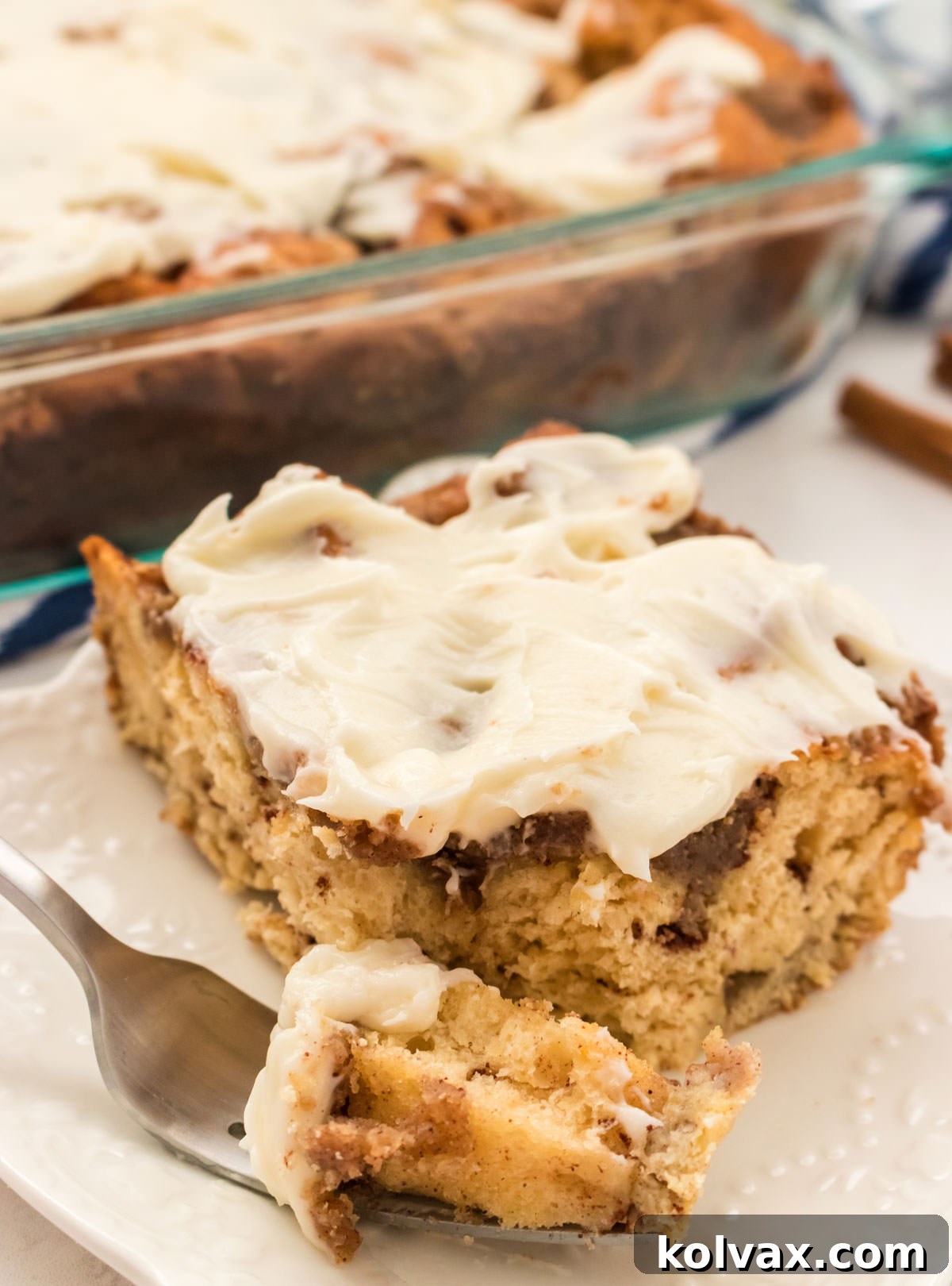 Close up of a piece of Cinnamon Roll Breakfast Casserole sitting on a white plate in front of the pan of remaining casserole.