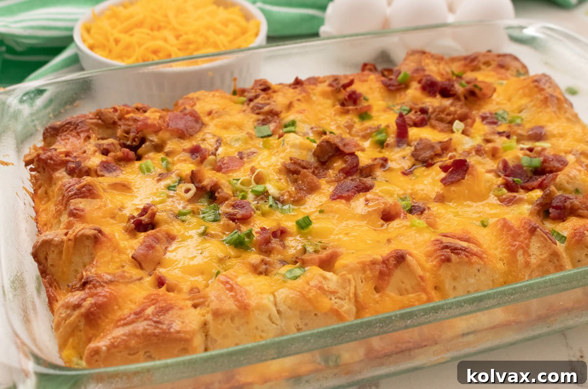 Close up of a golden-brown Bacon and Egg Breakfast Casserole in a glass baking pan, ready to serve, with a ramekin of cheese and a bowl of eggs in the background.