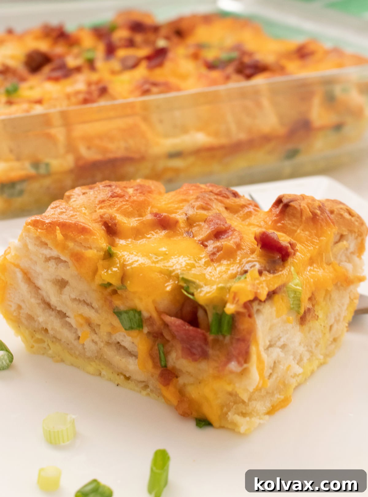 A close-up view of a perfectly baked slice of Bacon and Egg Breakfast Casserole on a white plate, with the rest of the casserole in the pan in the background.