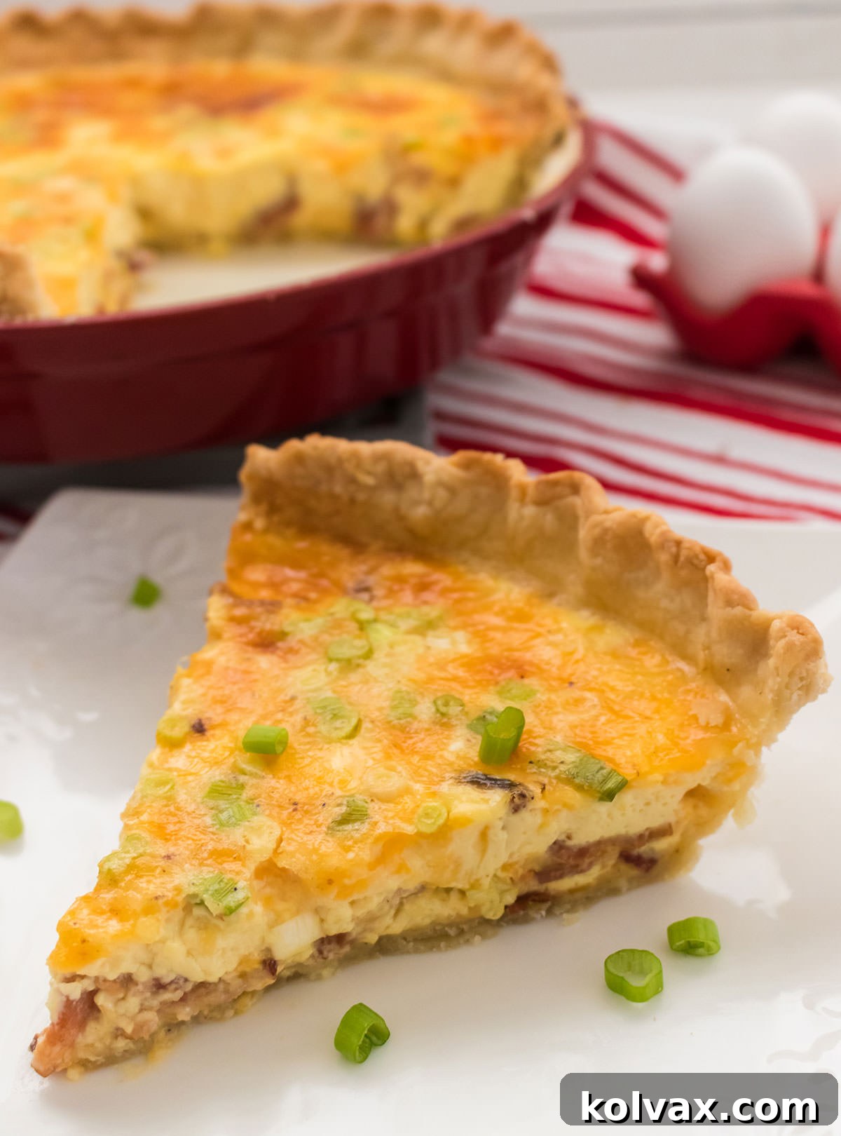 A delicious slice of golden-brown Quiche Lorraine placed on a crisp white plate, with the remainder of the quiche in its dish blurred in the background, ready for serving.