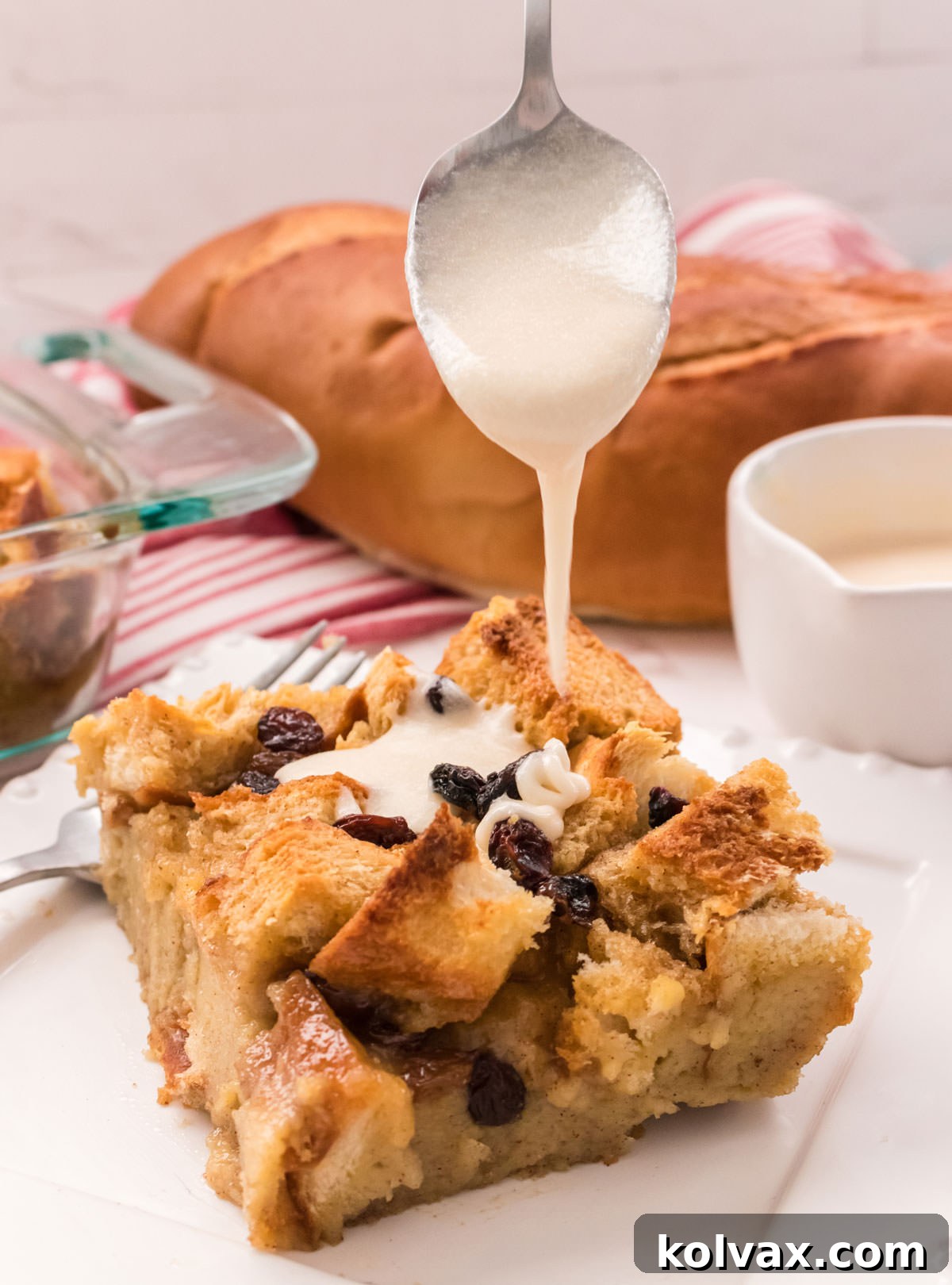 Close-up of a serving of warm bread pudding on a white plate, with a spoon drizzling rich vanilla sauce over the top, highlighting its appealing texture.