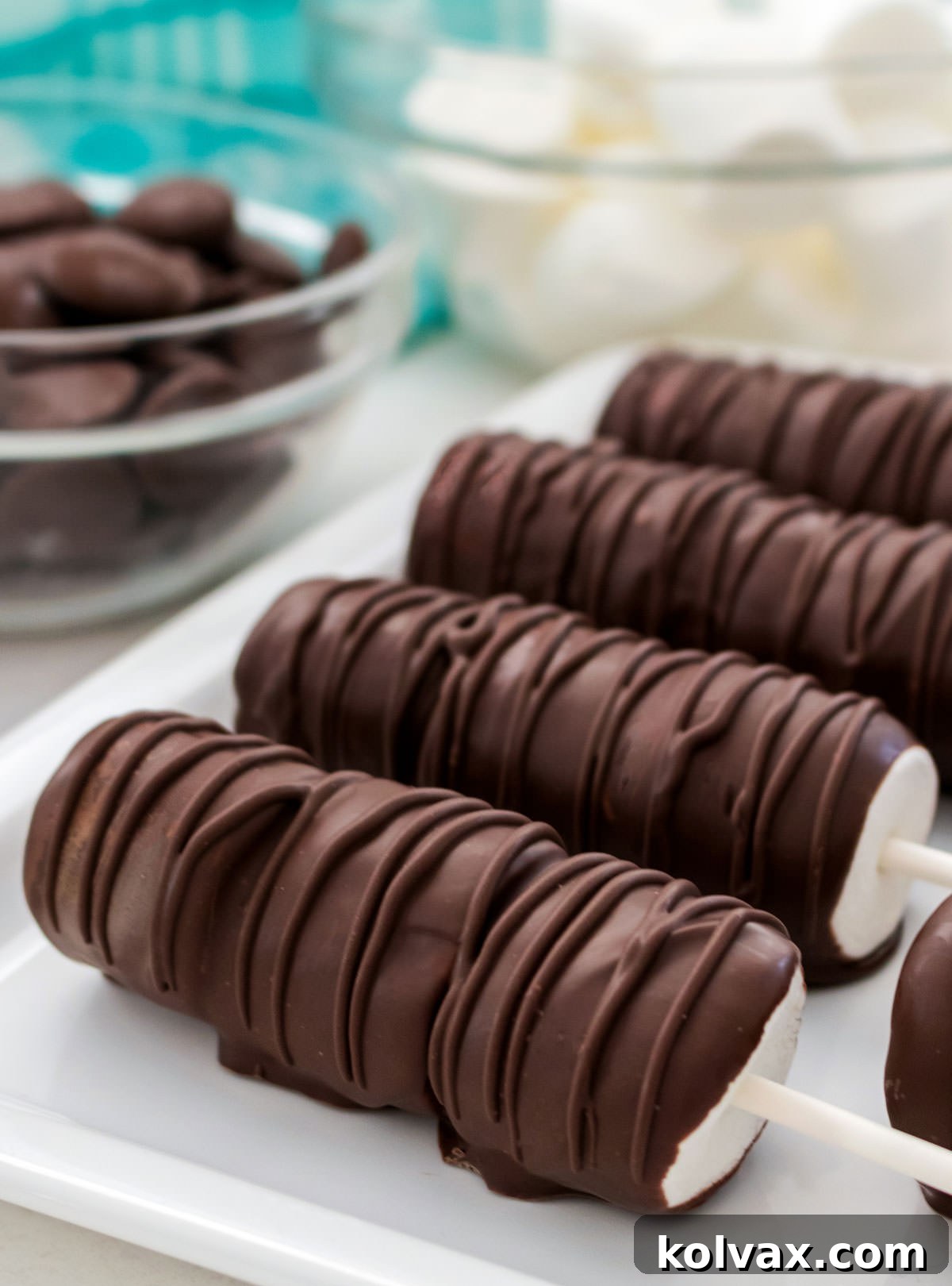 A beautiful close-up on a white platter featuring four perfectly crafted Chocolate Marshmallow Pops, elegantly displayed in front of bowls filled with melting chocolate and fresh marshmallows.