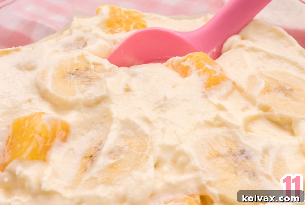 Closeup shot of Pineapple Banana Fluff in a glass bowl, showcasing pineapple and banana slices visible within the creamy fluff, with a pink spatula resting nearby.