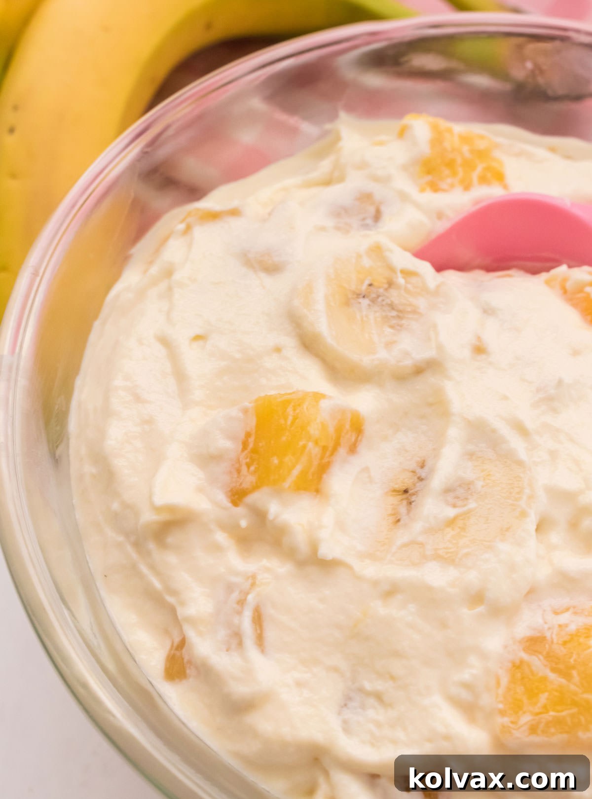 Closeup on a glass bowl filled with Pineapple Banana Fluff, perfectly chilled, with a whole banana and a pink table linen subtly blurred in the background, ready to be served.