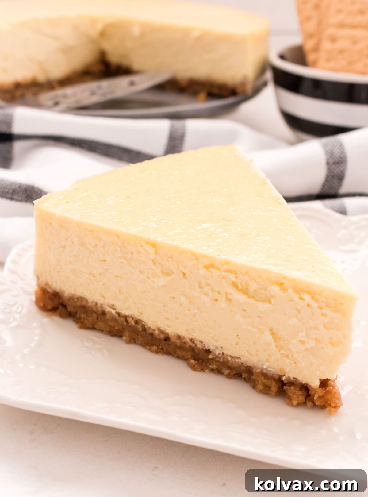 Closeup of a single piece of Classic Cheesecake sitting on a white plate in front of the remaining cheesecake in the background, ready to be enjoyed.