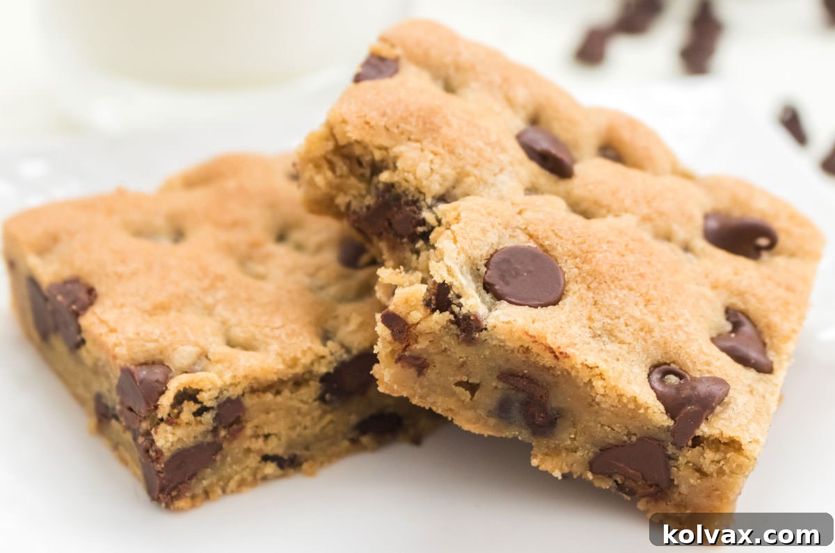 Two golden brown Chocolate Chip Cookie Bars sitting on a small white plate, elegantly placed in front of a glass of refreshing milk, surrounded by scattered chocolate chips.