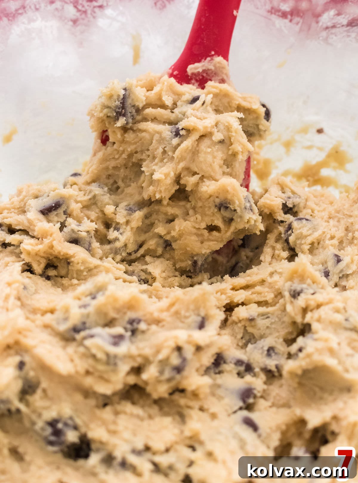 A clear glass mixing bowl filled with rich, ready-to-bake Chocolate Chip Cookie dough, with a vibrant red spatula resting inside, indicating completion of the mixing process.
