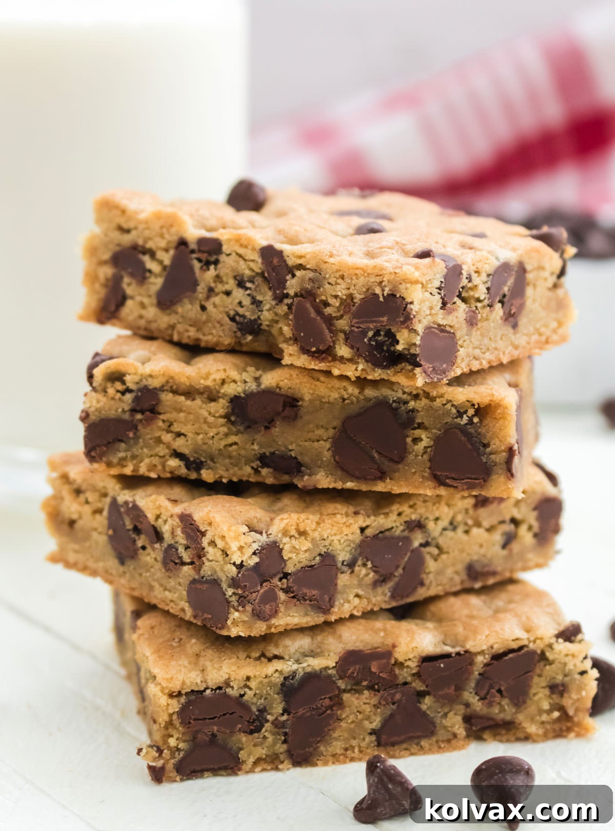 A delightful stack of four golden-brown Chocolate Chip Cookie Bars artfully arranged on a pristine white surface, positioned in front of a refreshing glass of milk and a warm red table linen, creating a cozy and inviting scene.