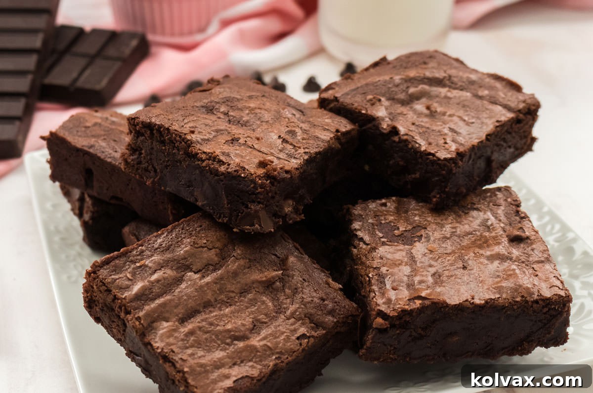 Close up of a plate of Homemade Fudge Brownies in front of a chocolate bar, a glass of milk and a pink table linen, highlighting their rich, dark texture.