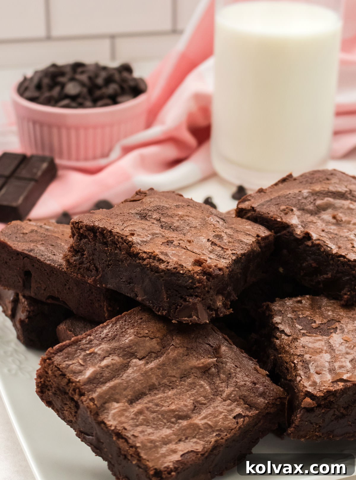 Close up on a white plate filled with Homemade Fudge Brownies along with a glass of milk and a ramekin of chocolate chips in the background, showcasing their irresistible texture.
