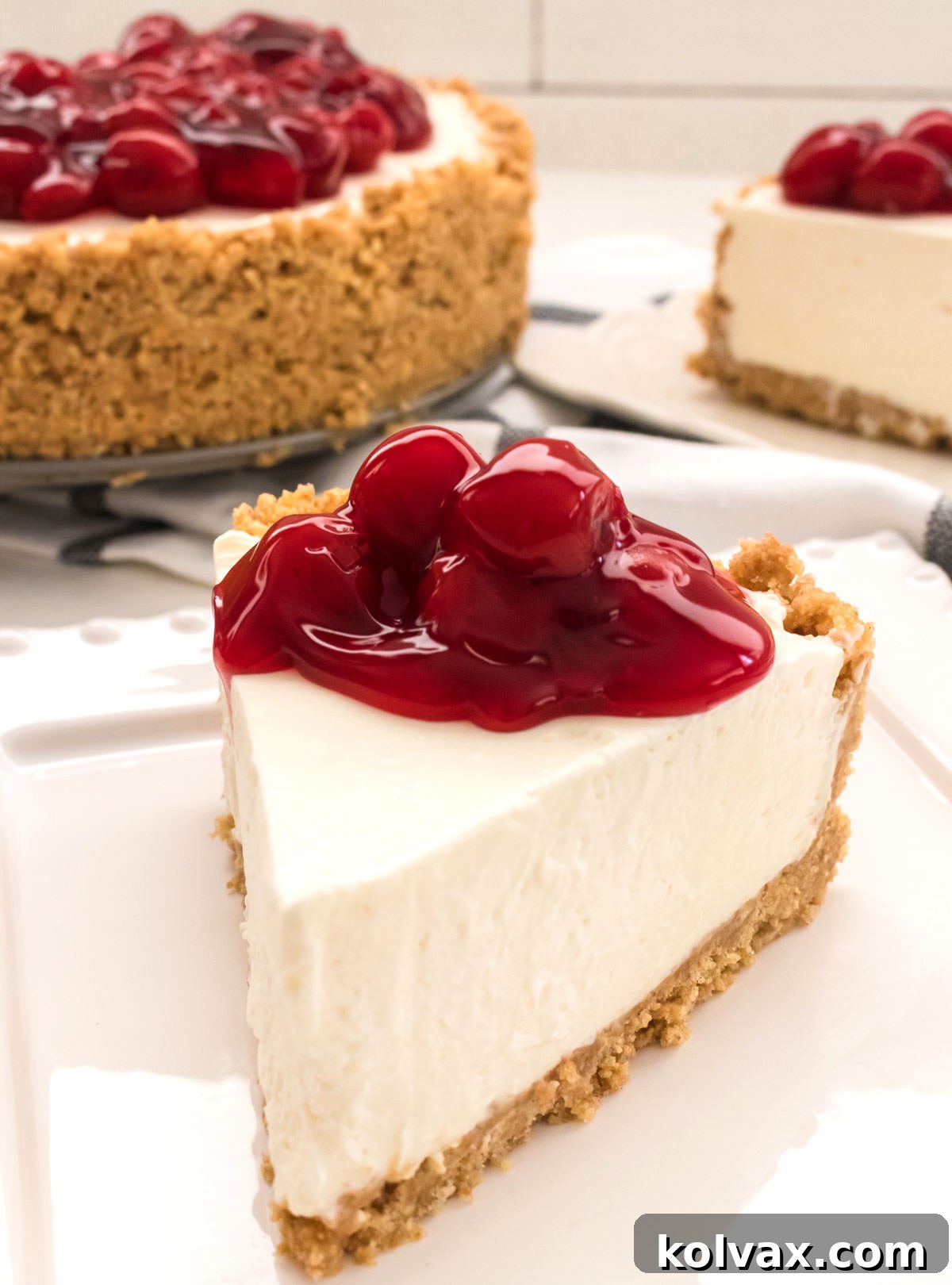 Close up of a single perfectly sliced piece of No Bake Cheesecake sitting on a clean white plate, with the rest of the beautifully set cheesecake visible in the blurred background.