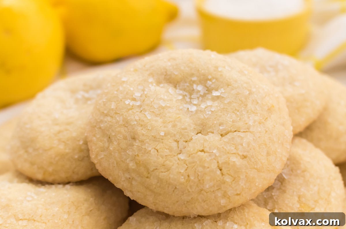 A beautiful stack of freshly baked Lemon Sugar Cookies, gleaming with sparkling sugar, sits proudly in front of a scattering of fresh lemons and a vibrant yellow ramekin. These cookies boast a perfect golden-brown hue, hinting at their soft, chewy interior and subtly crisp edges. Ideal for a refreshing spring or summer treat.