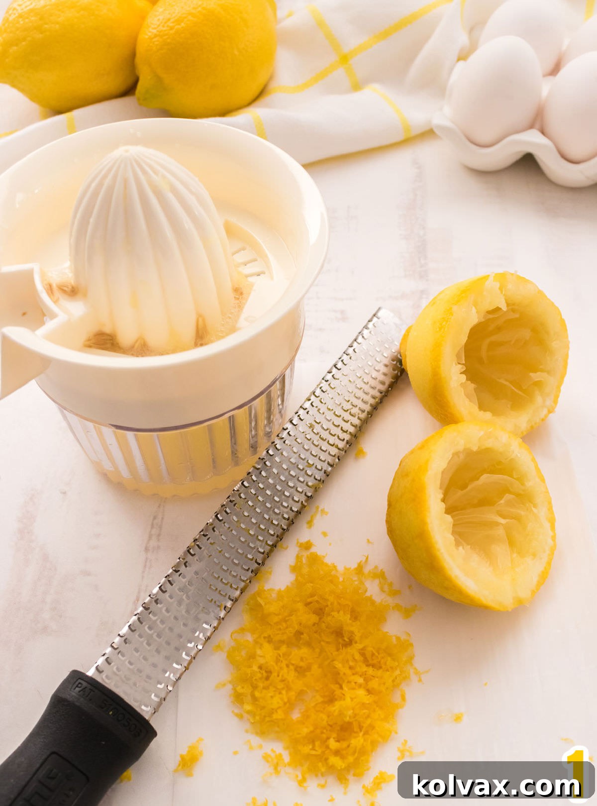 A close-up shot showcasing the essentials for infusing vibrant lemon flavor: a bright yellow lemon, a manual lemon juicer ready for action, and a Microplane zester covered in fragrant lemon zest. In the softly blurred background, a kitchen towel and fresh eggs hint at the delightful baking process ahead.