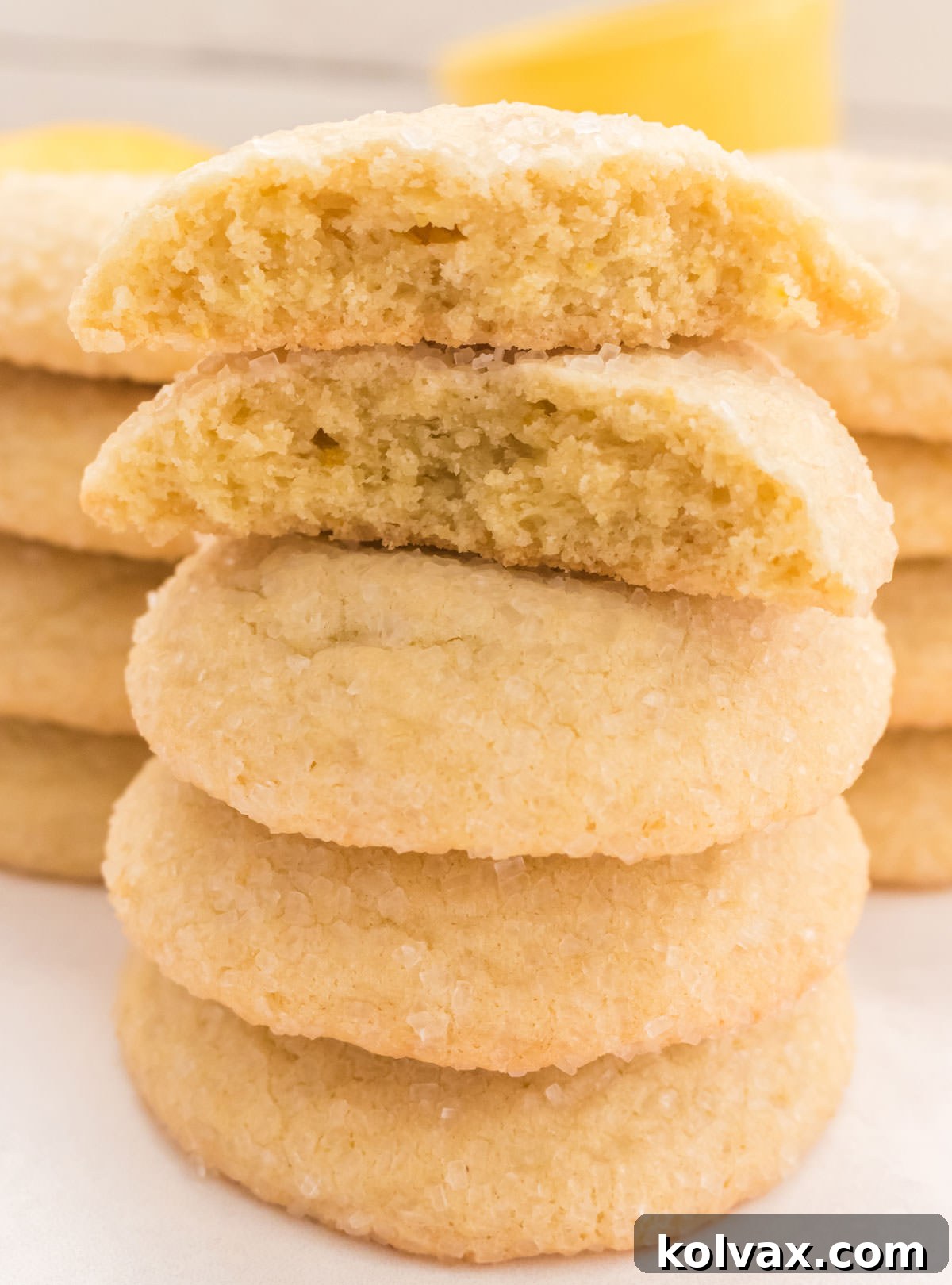 A stunning close-up of a perfectly arranged stack of Lemon Sugar Cookies, with additional stacks visible in the background. The top cookie is artfully broken in half, revealing its incredibly soft, chewy, and tender interior, while the edges maintain a delicate crispness. This image perfectly showcases the irresistible texture of these homemade lemon treats.