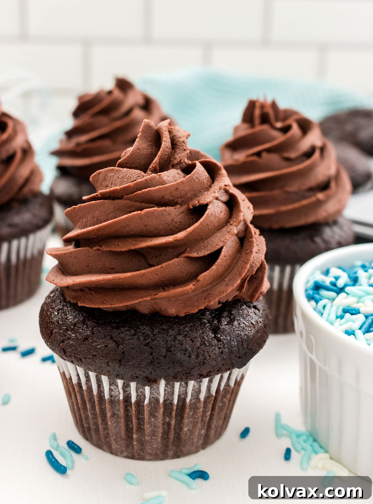 Close up on four Homemade Chocolate Cupcakes sitting on a white table surrounded by sprinkles and a blue table linen.