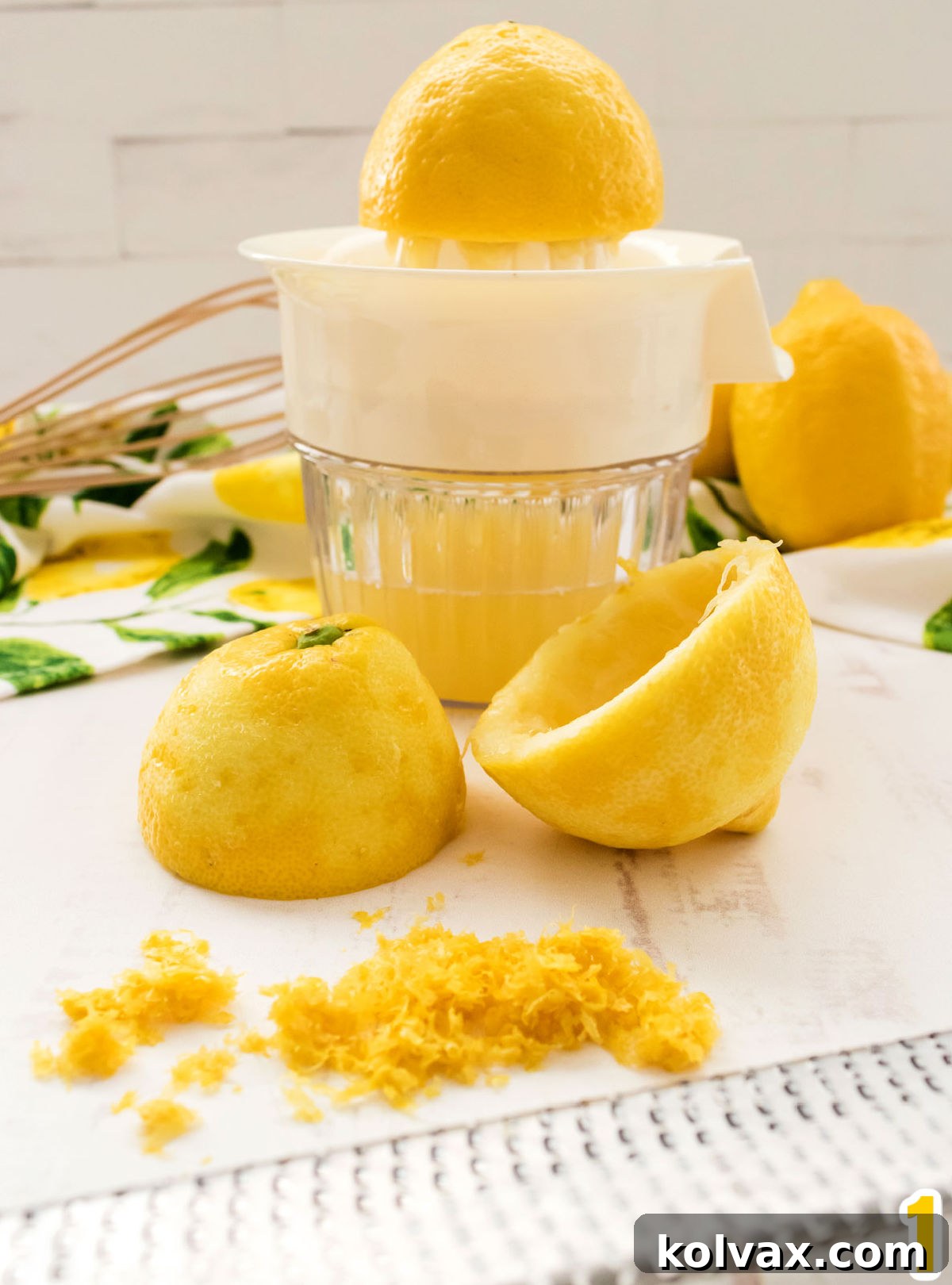 Close-up of a lemon juicer filled with fresh lemon juice, a microplane with lemon zest, a whisk, and whole lemons ready for preparation.