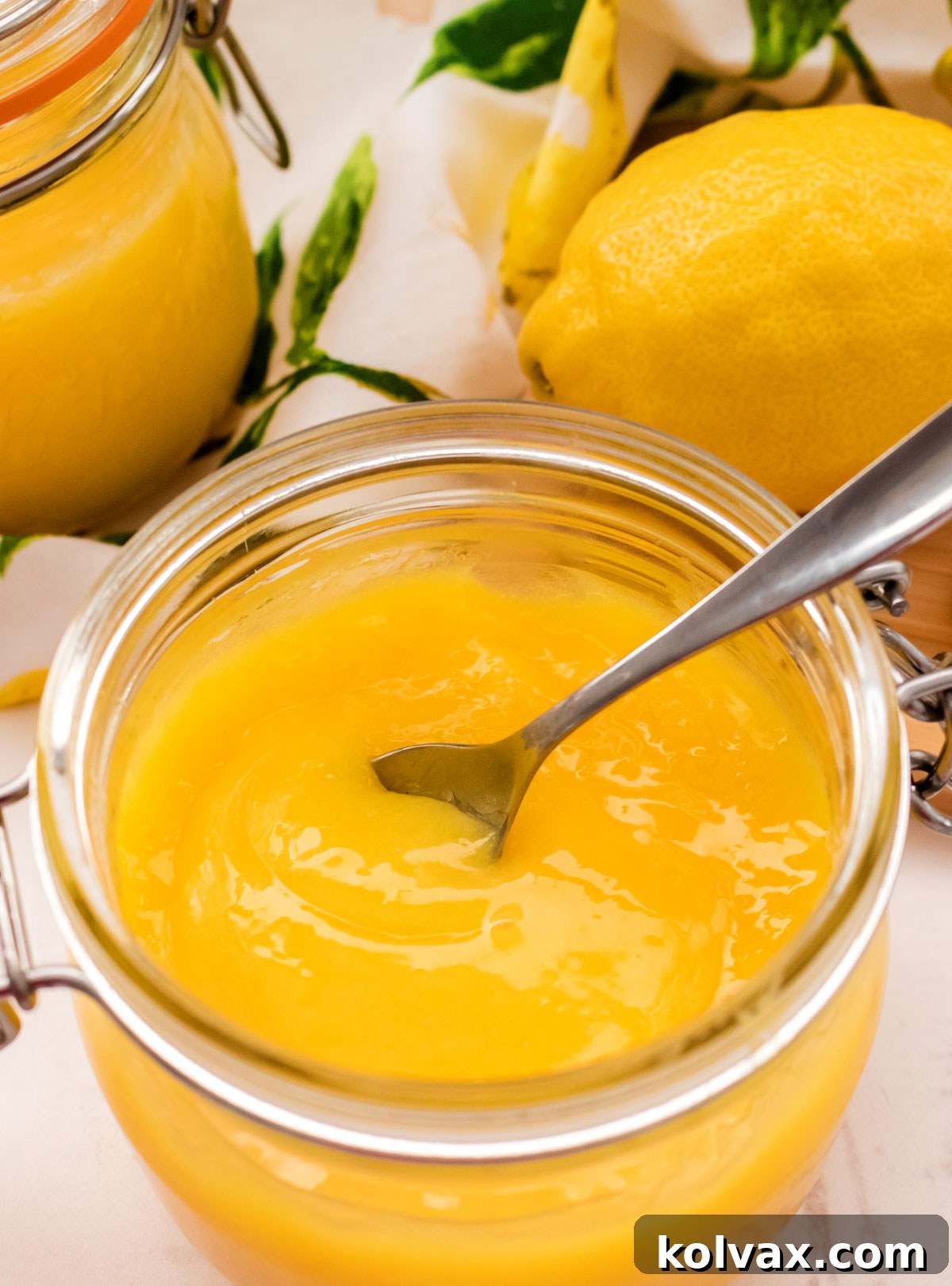 A charming scene featuring a glass jar filled with homemade Lemon Curd and a silver spoon, elegantly arranged with fresh lemons and a soft kitchen towel.