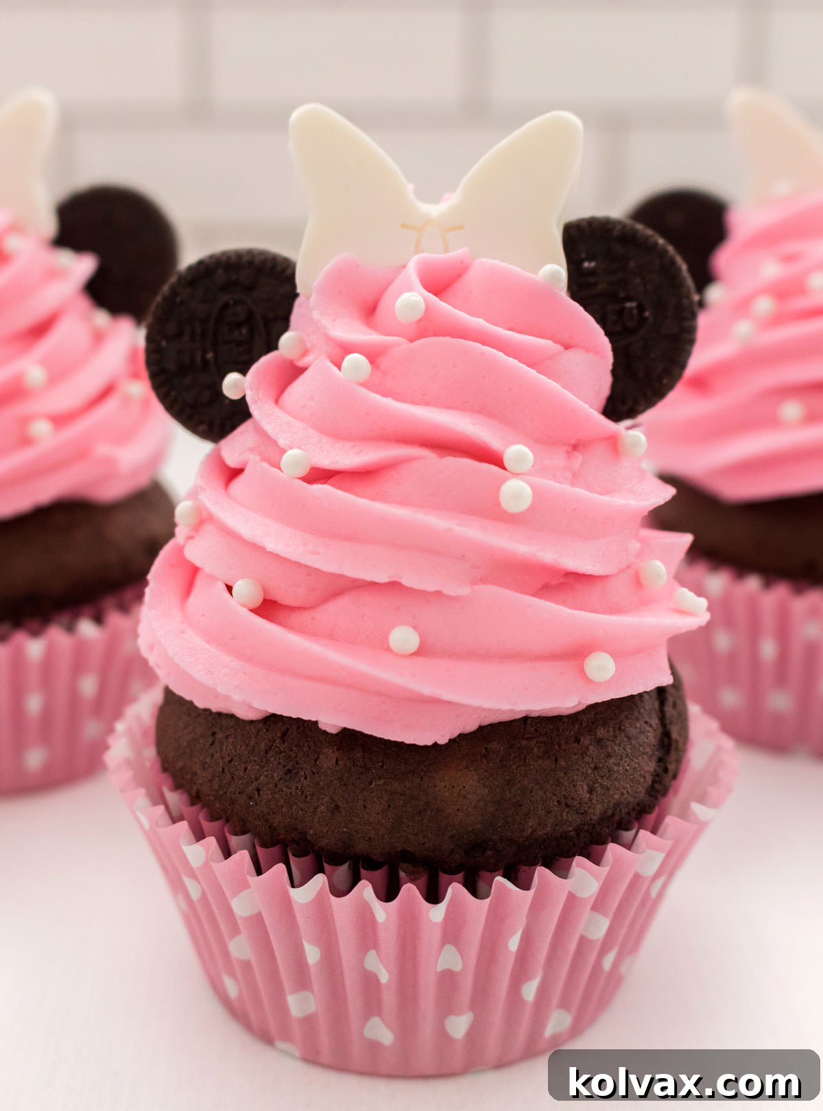 Close-up of a single beautifully decorated Minnie Mouse Cupcake with pink buttercream, Oreo ears, and a white chocolate bow, surrounded by other cupcakes.