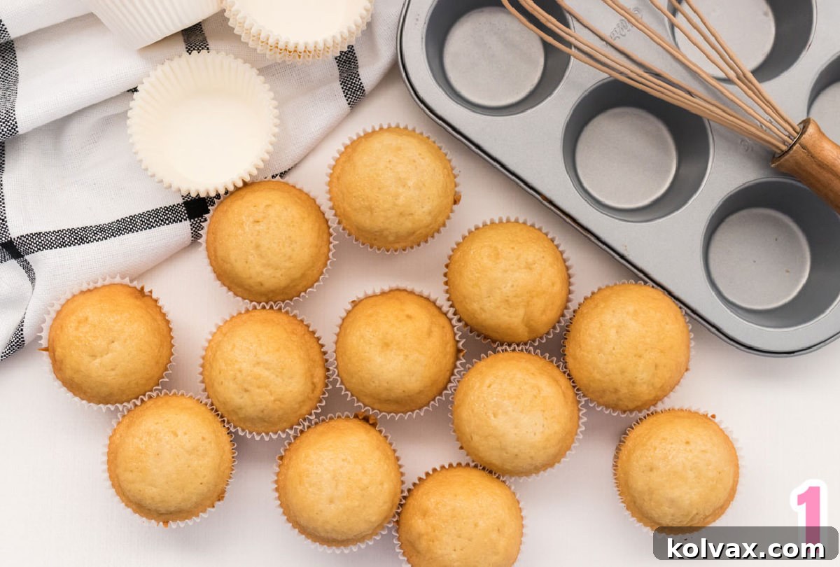 A close-up shot of a dozen freshly baked vanilla cupcakes, still in their pink liners, arranged on a pristine white table. A cupcake tin and a charming black and white kitchen towel are visible in the background.