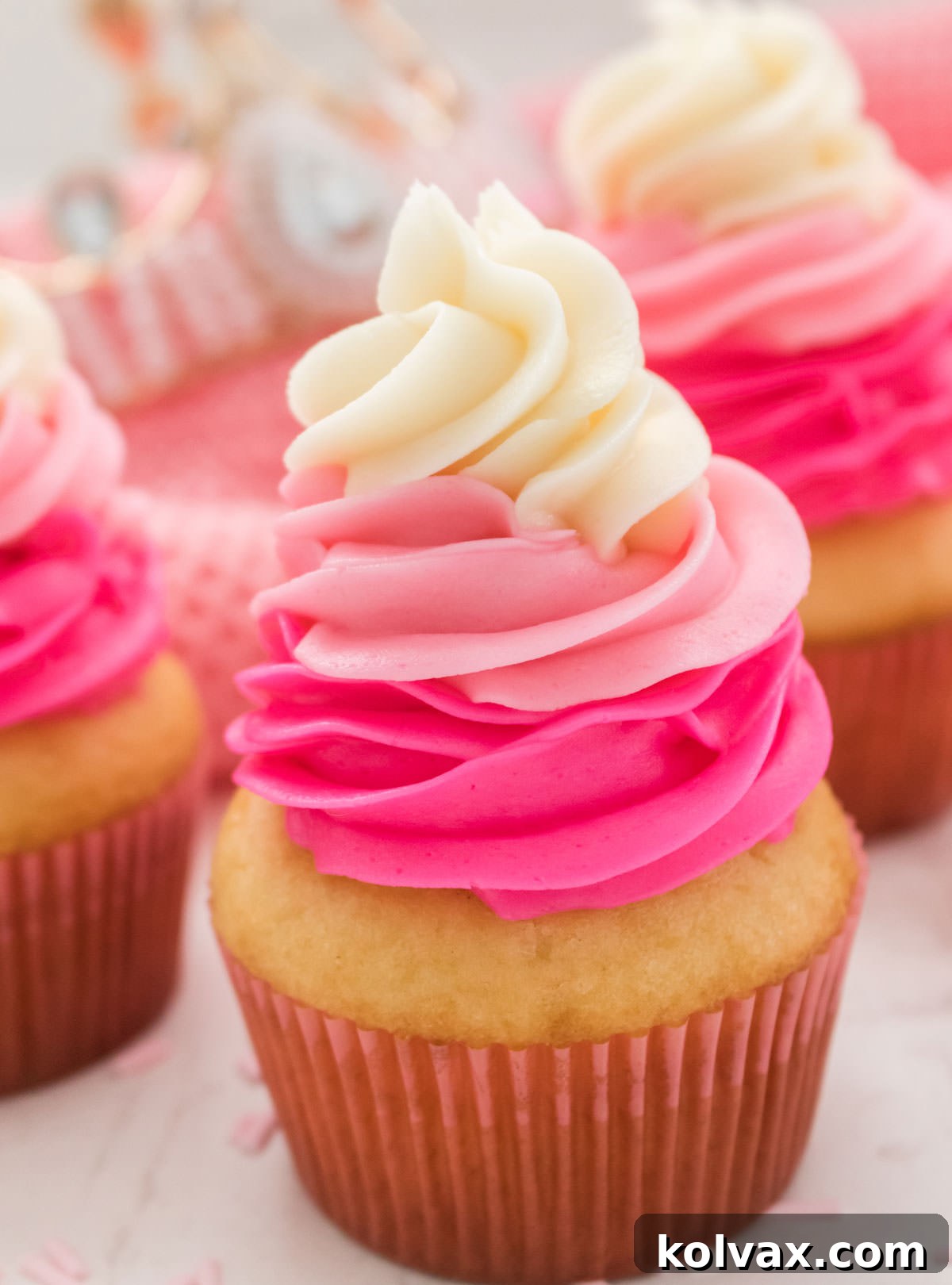 A stunning close-up of three beautifully frosted Princess Cupcakes with pink ombre swirls, elegantly displayed on a white table. A soft pink towel and a delicate pink princess tiara are visible in the background, completing the royal setting.