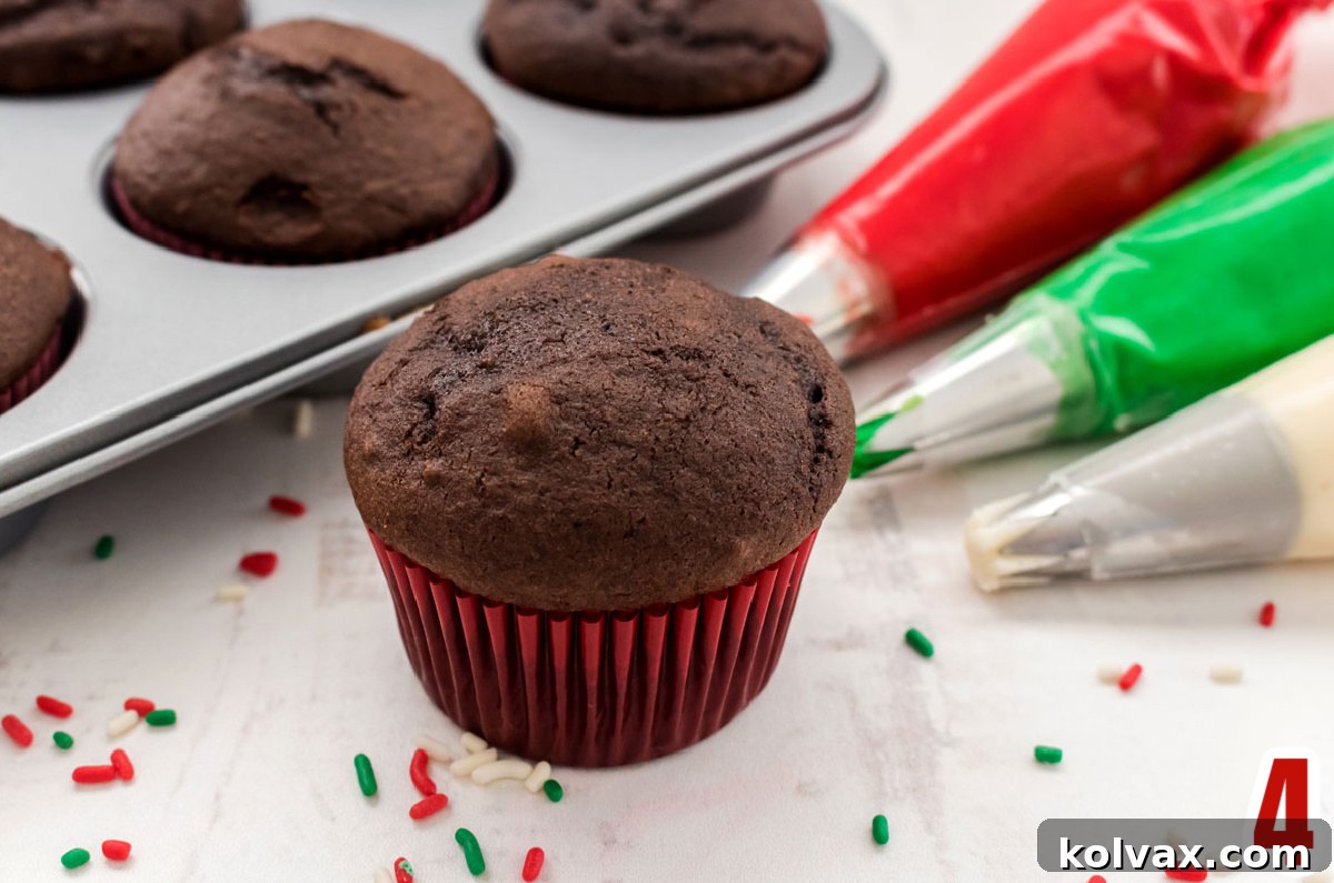 A chocolate cupcake in a vibrant red liner is positioned on a white table, next to three separate decorating bags, each filled with red, green, and white buttercream frosting and fitted with a piping tip, ready for the swirl.