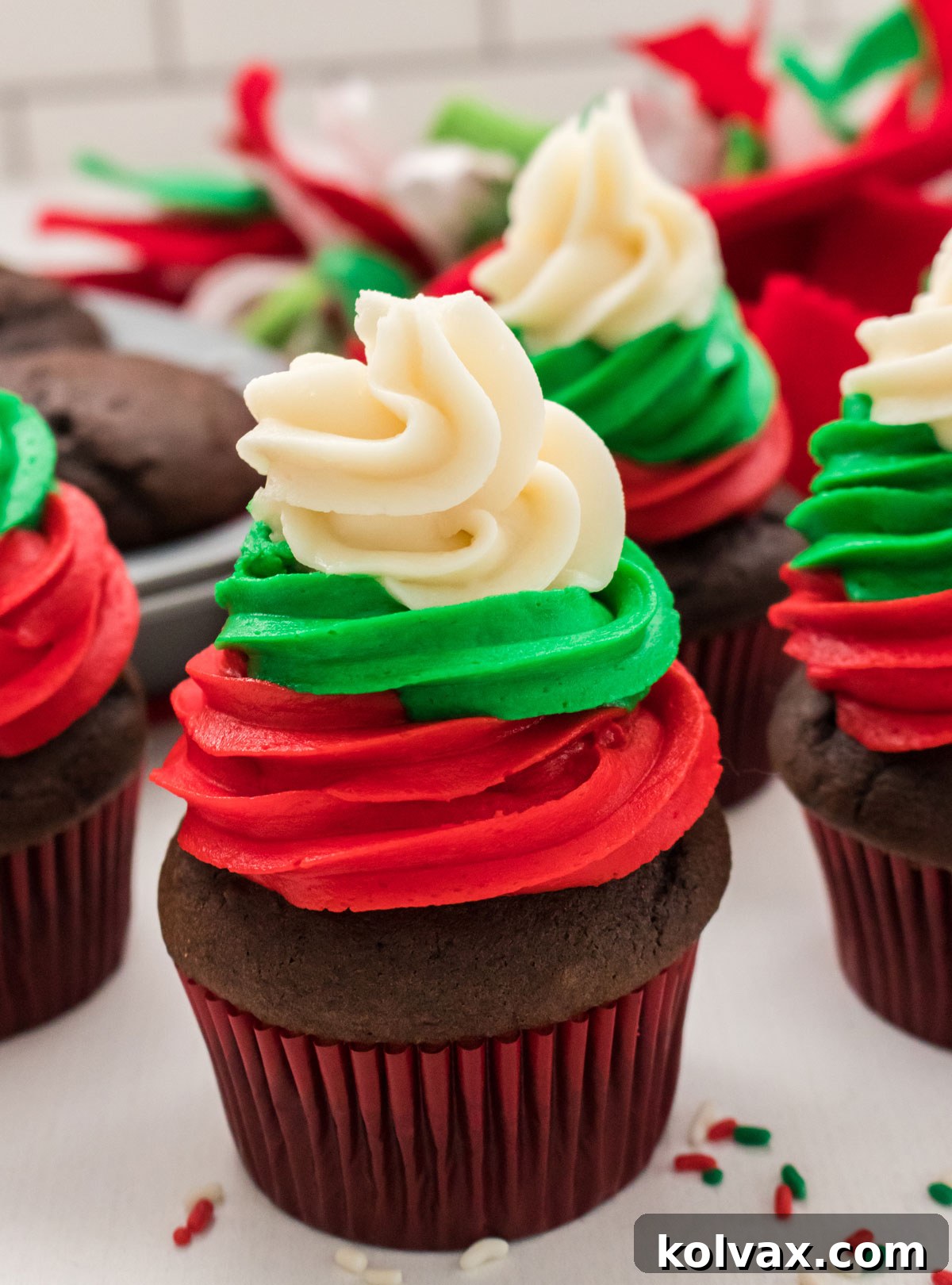 Closeup on four beautifully frosted Easy Christmas Cupcakes, adorned with red, green, and white swirls, sitting elegantly on a white table. In the background, a tin of un-frosted chocolate cupcakes and festive Christmas decorations complete the holiday scene.