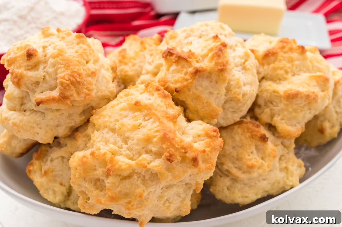 A beautiful white serving bowl brimming with golden Buttermilk Drop Biscuits, set against a warm red table linen, with a classic white butter dish nearby.