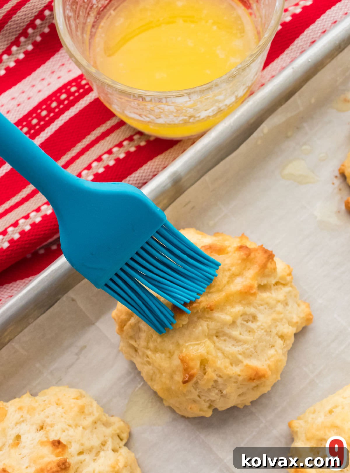 A close-up shot of a blue pastry brush delicately applying melted butter to the warm, freshly baked Buttermilk Drop Biscuits just out of the oven, enhancing their golden glow.