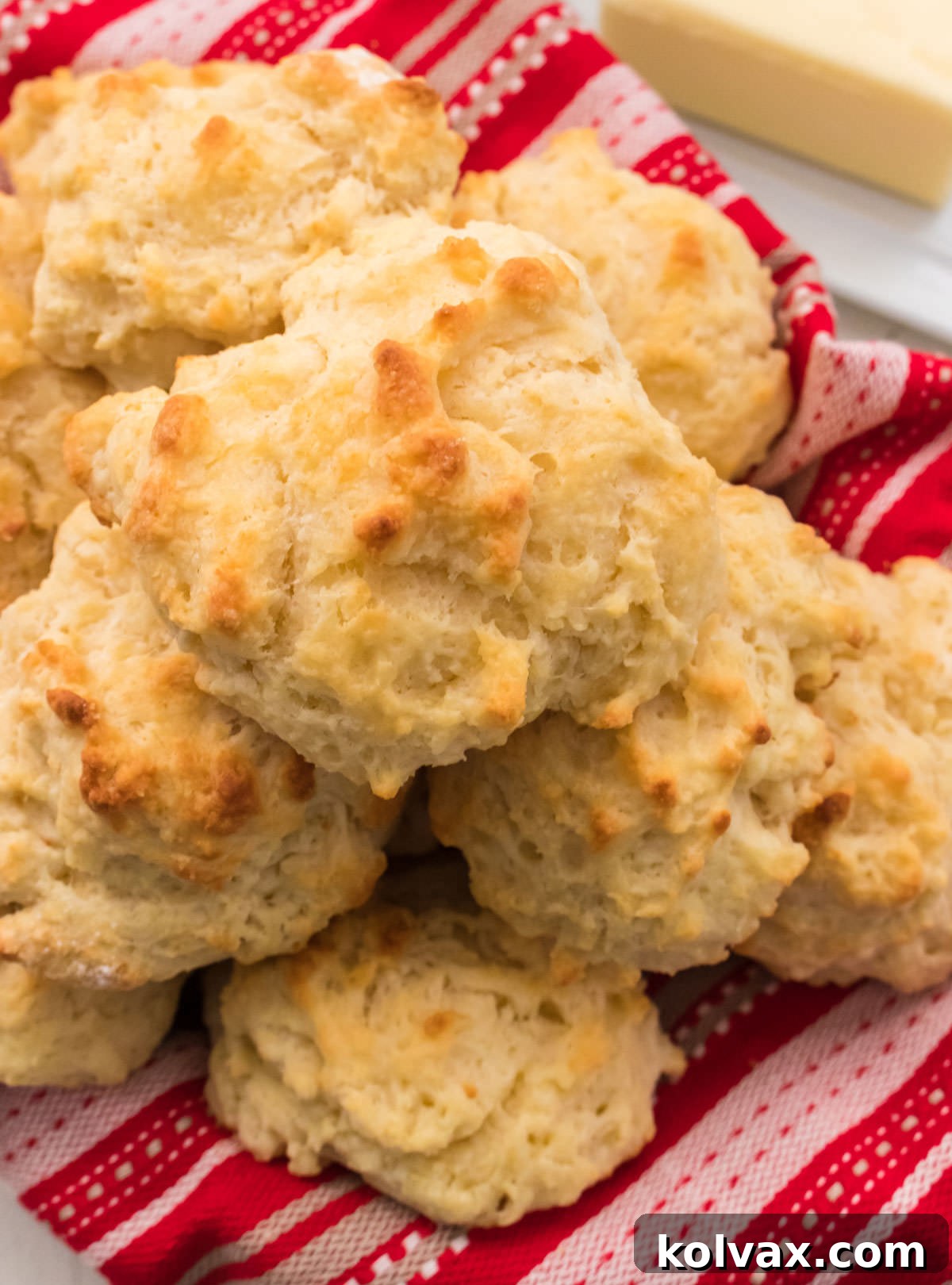 A rustic basket filled with a stack of golden Buttermilk Drop Biscuits, nestled beside a red checkered towel and a simple butter dish, ready to be enjoyed.