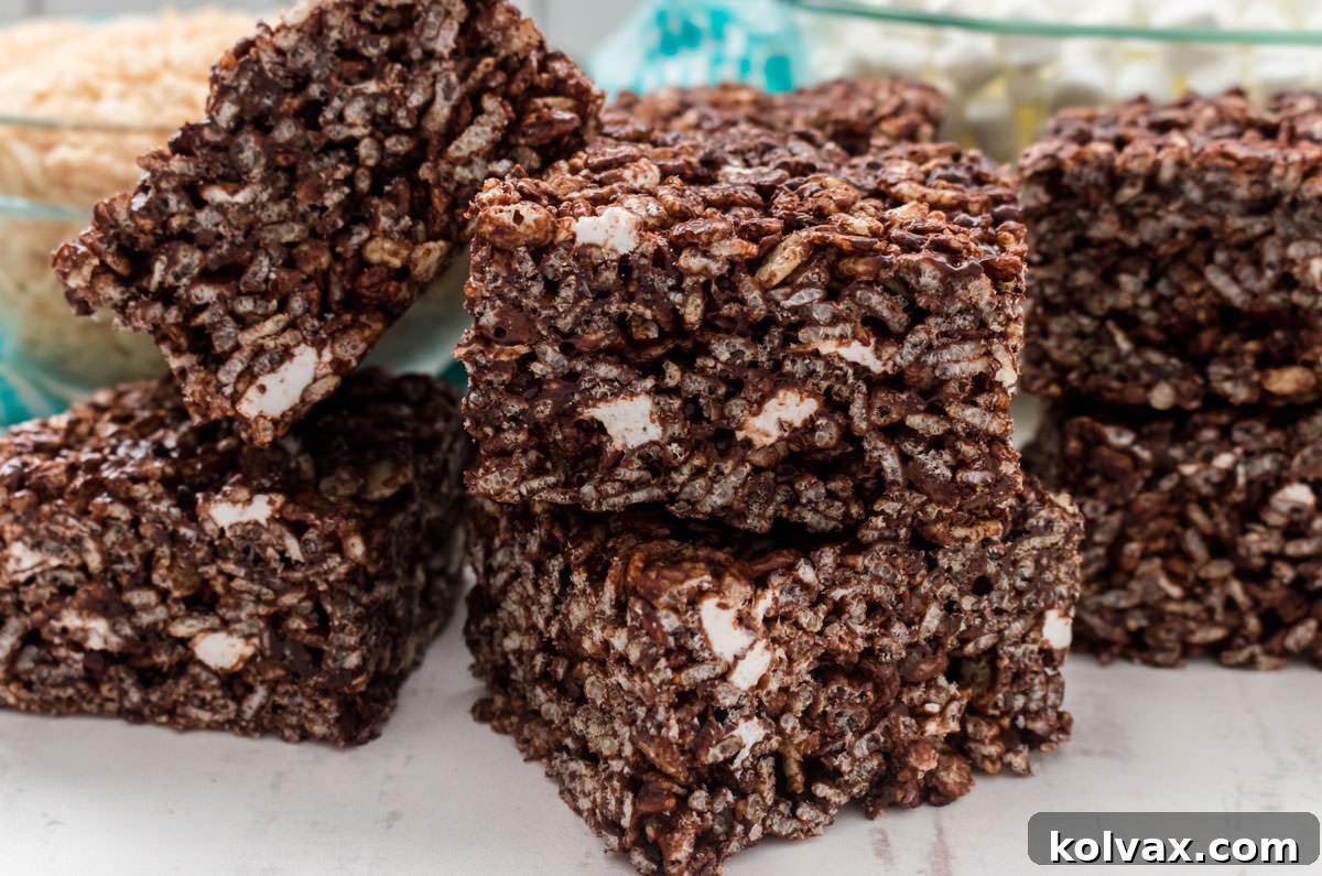 Closeup on a stack of six Chocolate Rice Krispie Treats sitting on a white table in front of bowls of cereal and marshmallows.