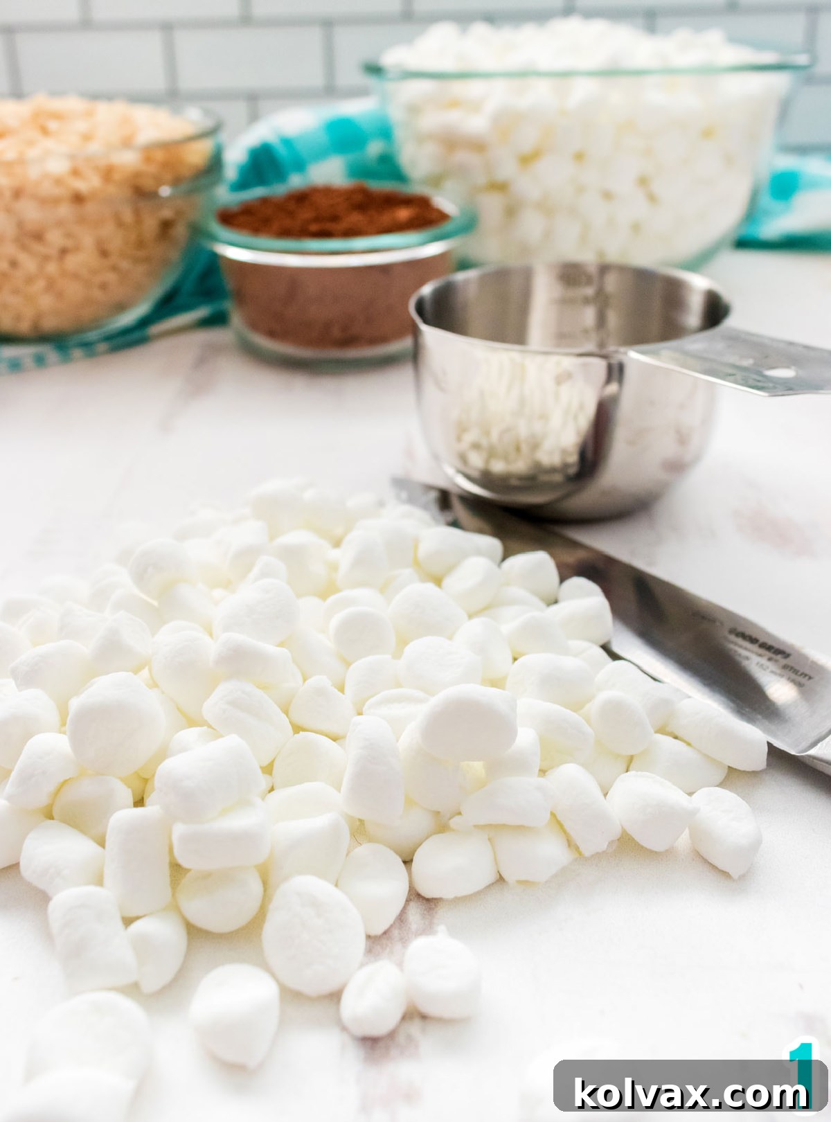 Chopped up Mini Marshmallows laying on a white table sitting next to a measuring cup, and bowls of cocoa powder, marshmallows and Rice Krispie Cereal.