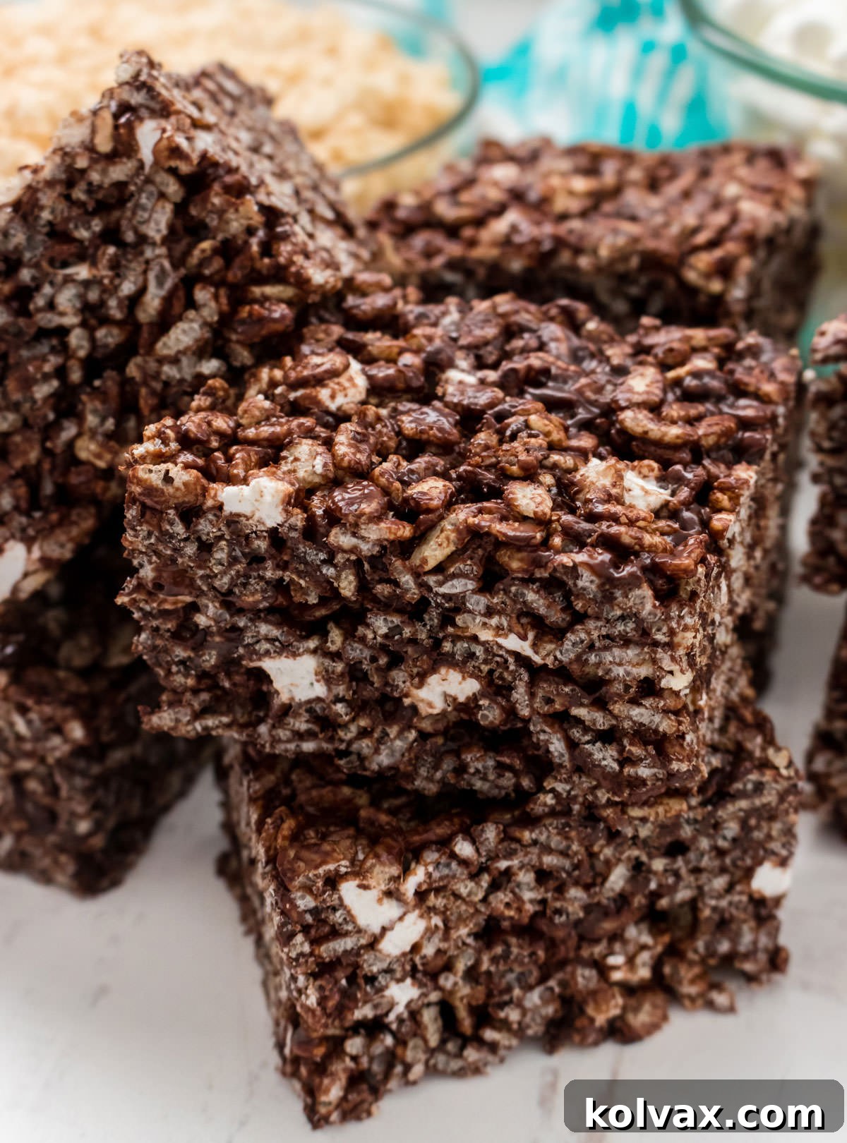 Closeup on a stack of Chocolate Rice Krispie Treats sitting on a white surface in front of a glass bowl filled with Rice Krispie Cereal.