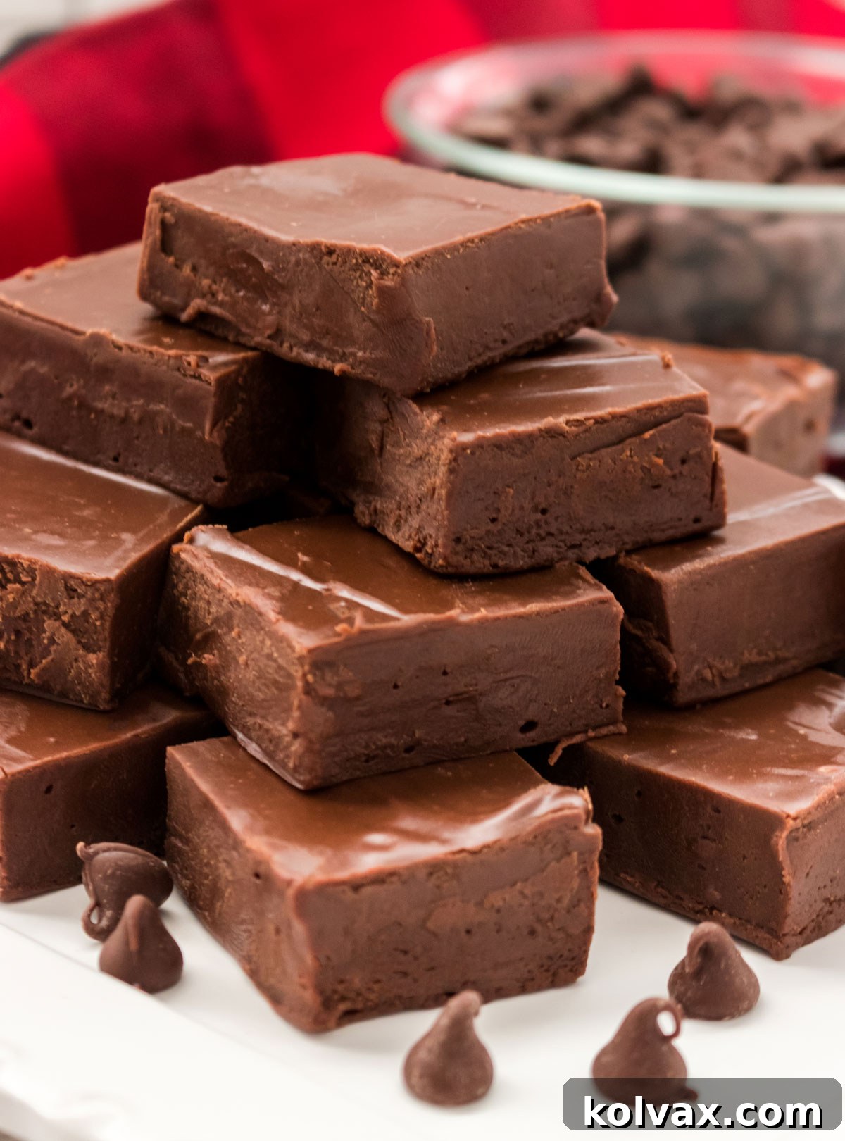 Closeup on a stack of Easy Chocolate Fudge sitting on a white surface in front of a red linen and a glass bowl filled with chocolate chips.