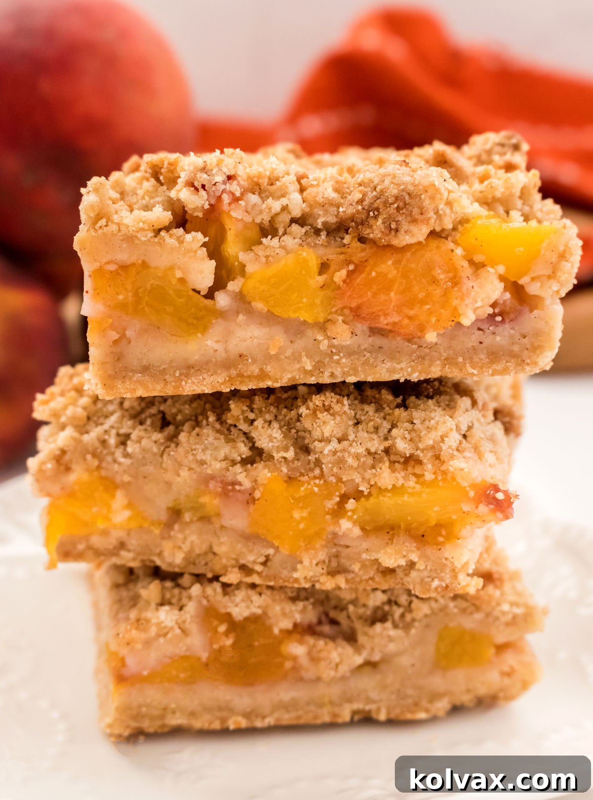 Closeup on a stack of Peach Crumble Bars sitting on a white plate in front of a orange towel and three fresh peaches.