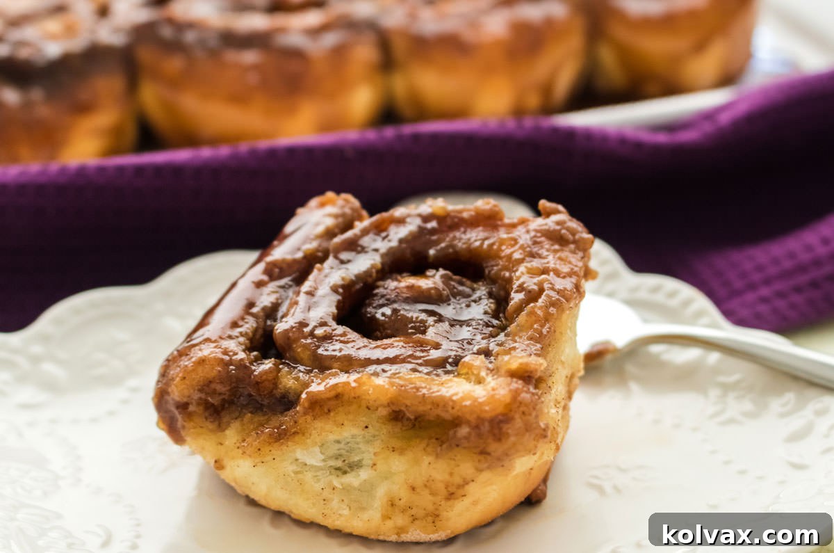 A single golden-brown Caramel Sticky Bun, glistening with caramel sauce, served on a pristine white plate, with a soft purple kitchen towel creating a cozy backdrop. The perfect holiday breakfast treat.
