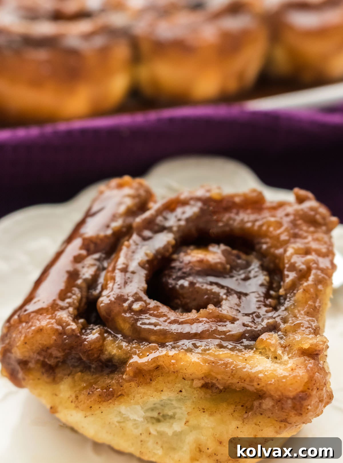 A close-up shot of a single Caramel Sticky Bun on a white plate, positioned in front of a larger platter brimming with the rest of the freshly baked pastries, showcasing their golden-brown color and glossy caramel topping.