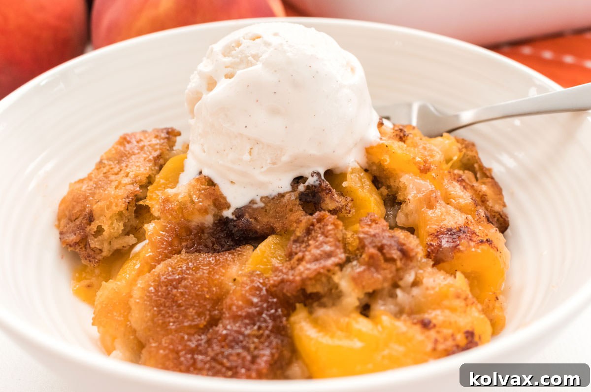 Heirloom Peach Cobbler 2 Closeup on a white bowl filled with warm Old Fashioned Peach Cobbler, topped with a generous scoop of melting vanilla ice cream, positioned in front of a rustic stack of fresh, ripe peaches.