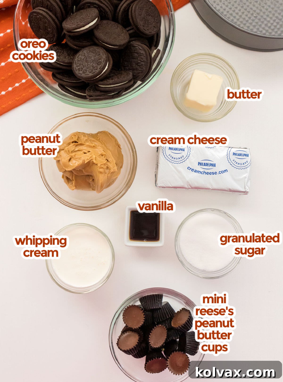 All the necessary ingredients laid out for making No-Bake Peanut Butter Cheesecake, including Oreos, melted butter, creamy peanut butter, blocks of cream cheese, granulated sugar, vanilla extract, heavy whipping cream, and mini Reese's Peanut Butter Cups.
