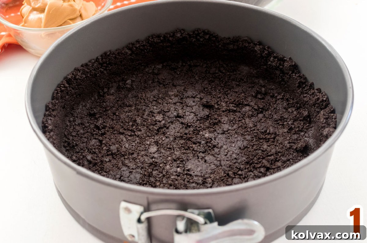 A meticulously pressed Oreo Cookie crust lining a silver springform pan, sitting ready on a clean white surface, awaiting its decadent filling.