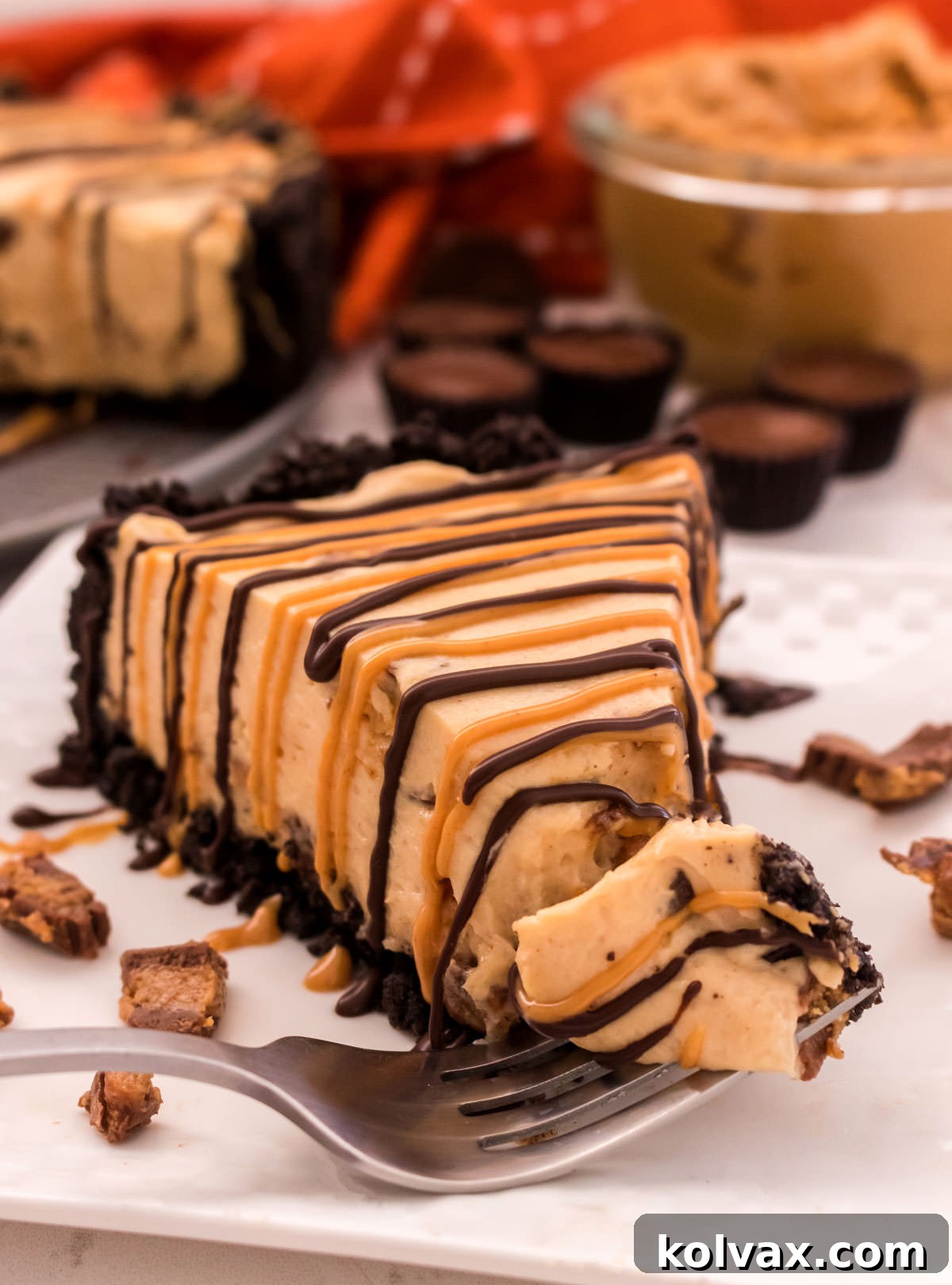 A single, impeccably cut slice of No-Bake Peanut Butter Cheesecake rests on a pristine white plate. In the background, the remaining cheesecake, a bowl of creamy peanut butter, and a scattering of Mini Reese's Peanut Butter Cups hint at the deliciousness.