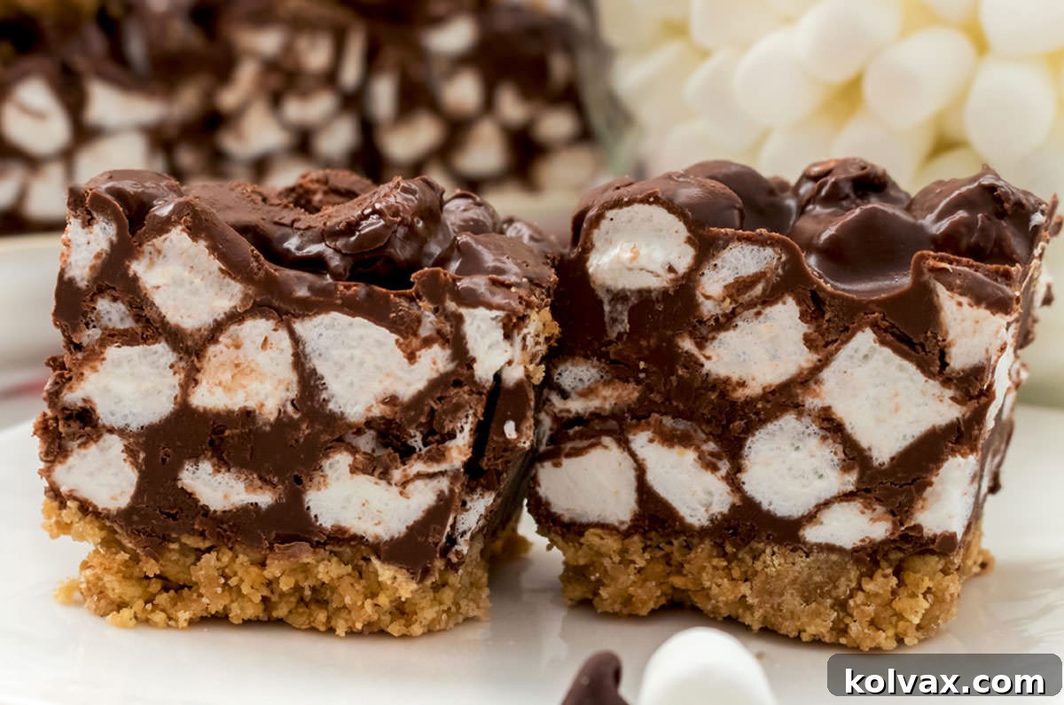 Closeup on two Chocolate Marshmallow Bars sitting on a white table in front of a bowl of mini marshmallows, showcasing their delightful layers.