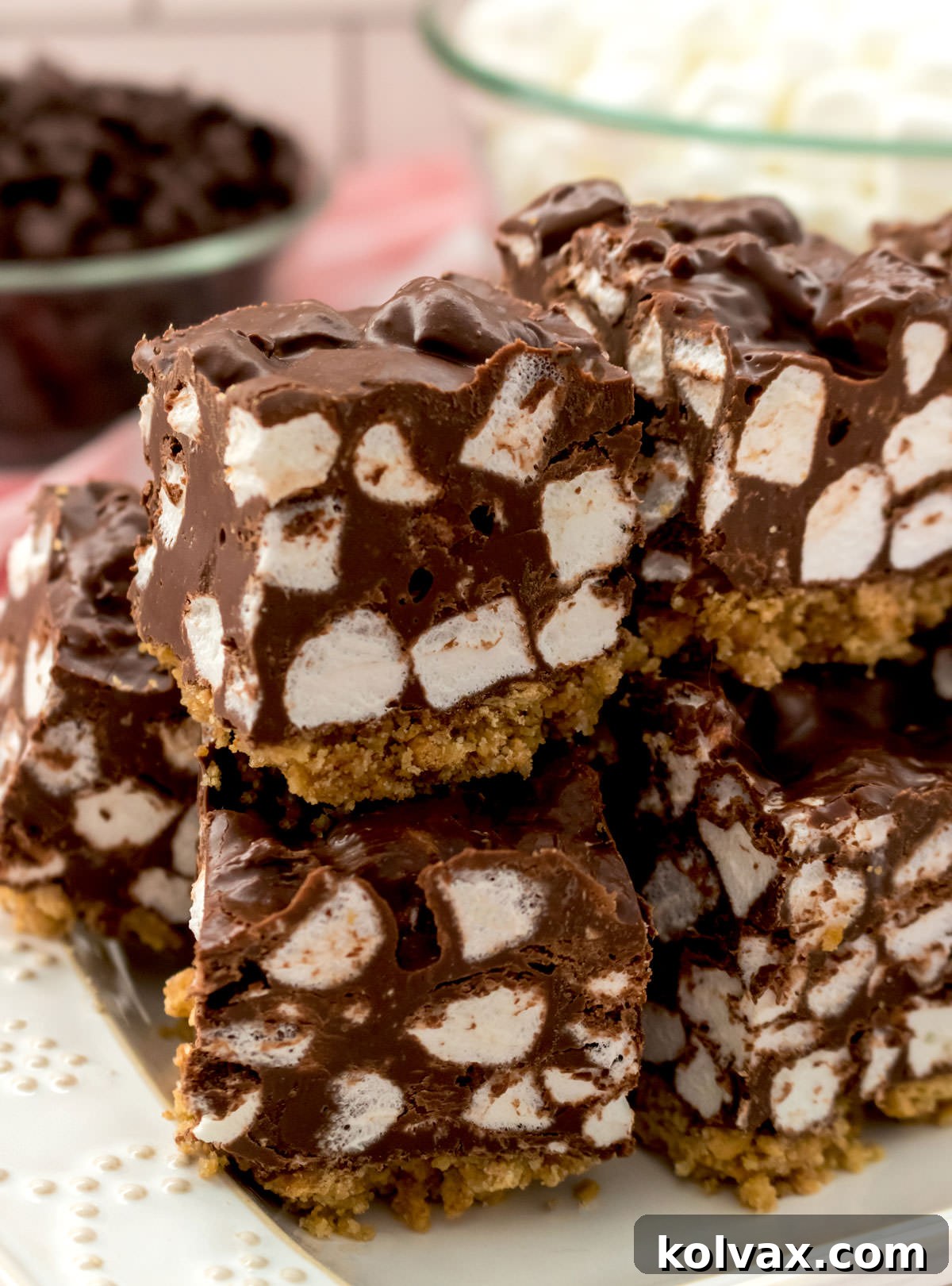 A closeup of a stack of Chocolate Marshmallow Bars presented on a white serving platter, with glass bowls of chocolate chips and mini marshmallows in the background.