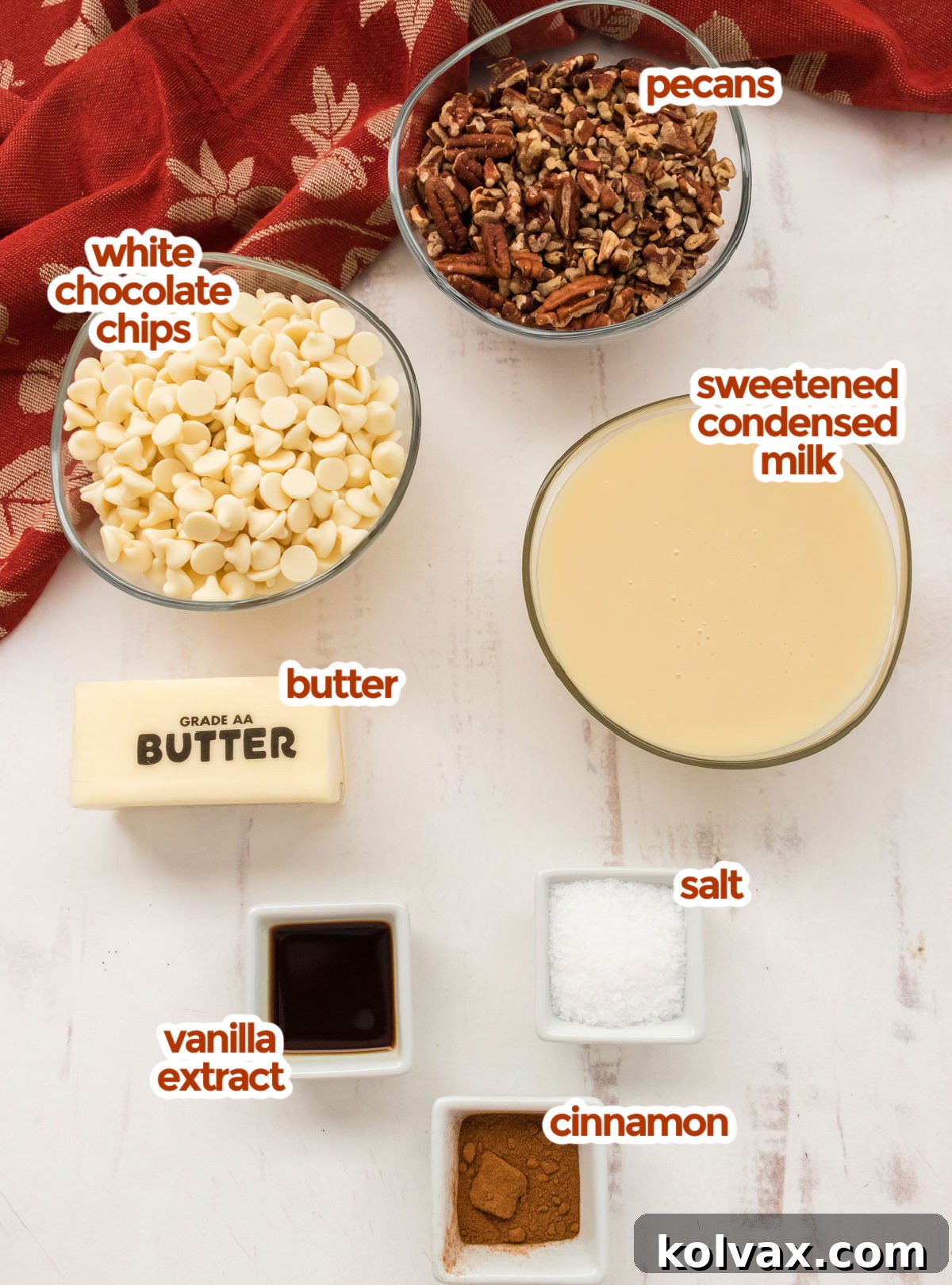 All the essential ingredients for Butter Pecan Fudge laid out on a white surface: pecans, sweetened condensed milk, white chocolate chips, butter, vanilla extract, cinnamon, and a pinch of salt.