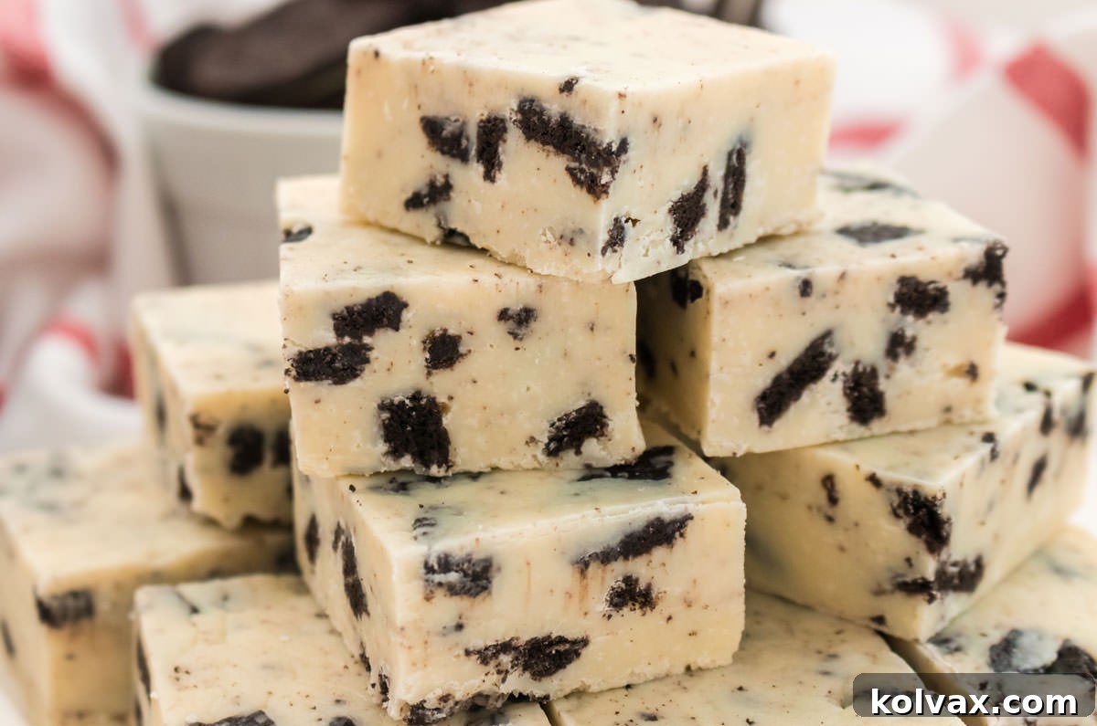 Closeup on a stack of homemade Oreo Fudge, showcasing its rich white chocolate base and abundant Oreo cookie chunks. The fudge pieces are arranged artfully in front of a textured kitchen towel and a white bowl overflowing with whole Oreo Cookies, inviting a delicious tasting experience.