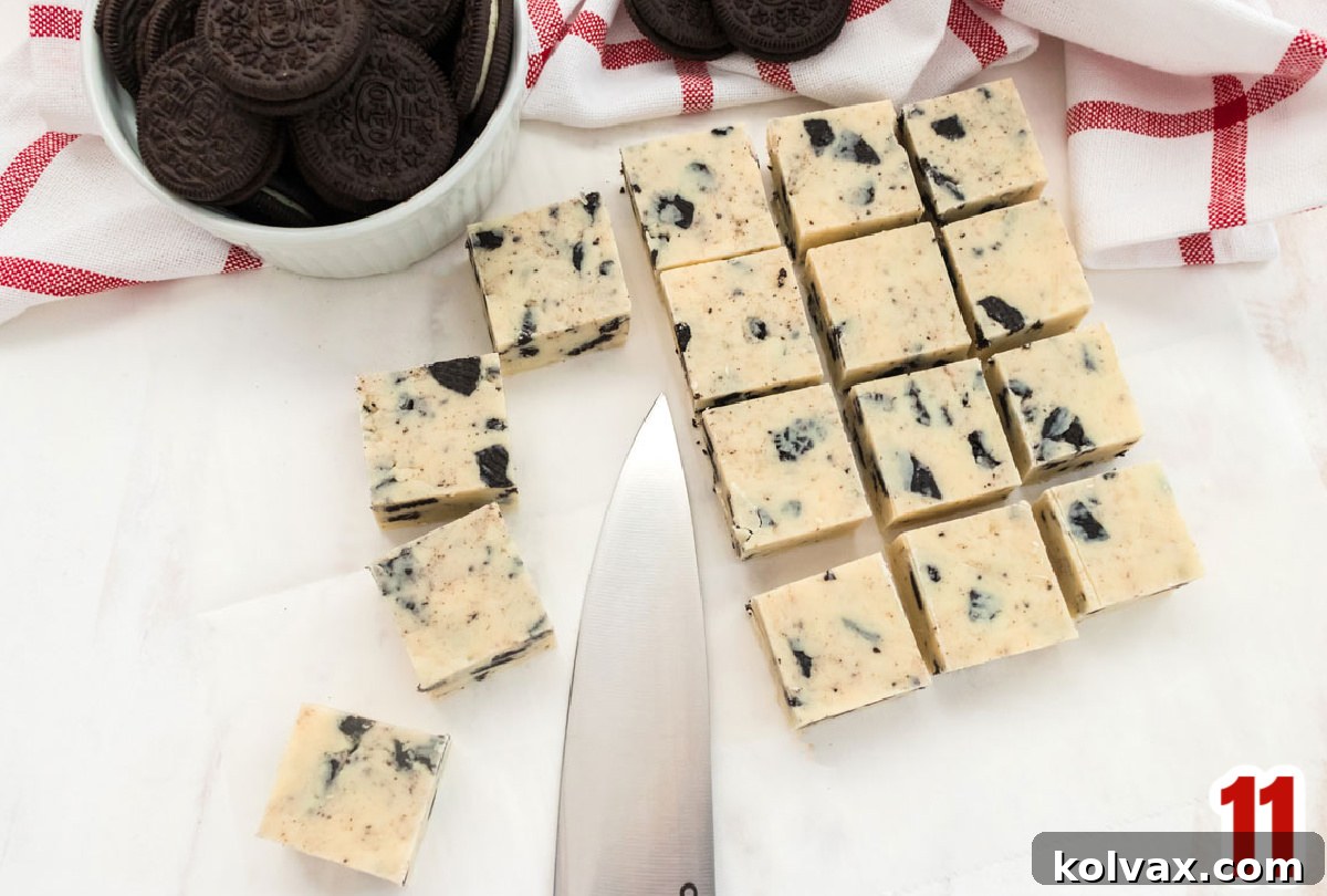 Sixteen perfectly cut pieces of Oreo Fudge arranged in neat rows on a white surface, accompanied by a sharp knife and a small ramekin brimming with whole Oreo Cookies, ready for enjoyment.