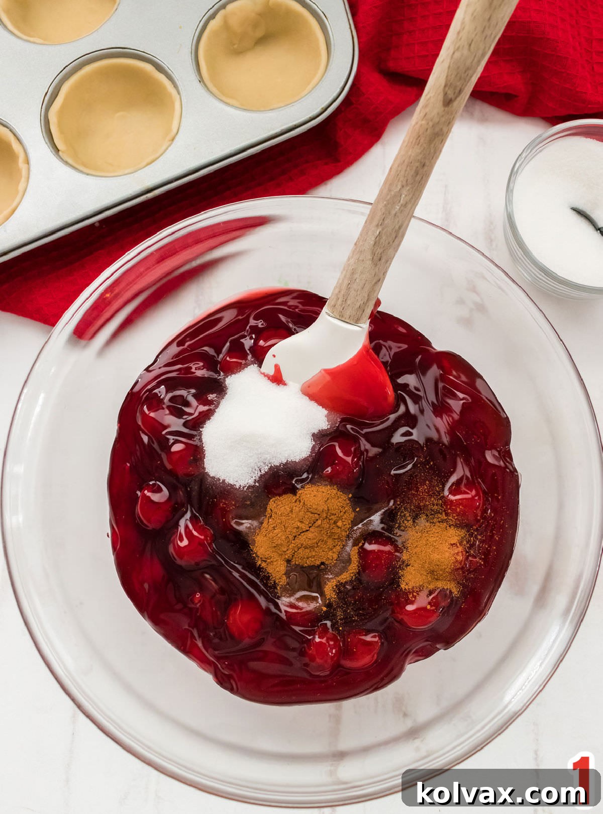 Bite-Sized Cherry Delights 4 A close-up of a glass bowl filled with vibrant red canned cherry pie filling, with granulated sugar, ground cinnamon, and a small bottle of almond extract waiting to be mixed in. The scene captures the anticipation of enhancing the filling's flavor.